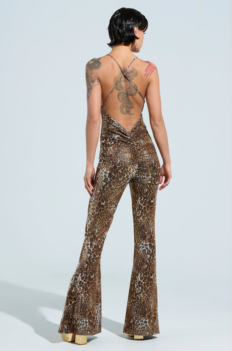 With short black hair and visible tattoos on one arm, a person stands confidently against a plain background, wearing the FEELIN FINE LEOPARD PRINT JUMPSUIT and rocking platform shoes.