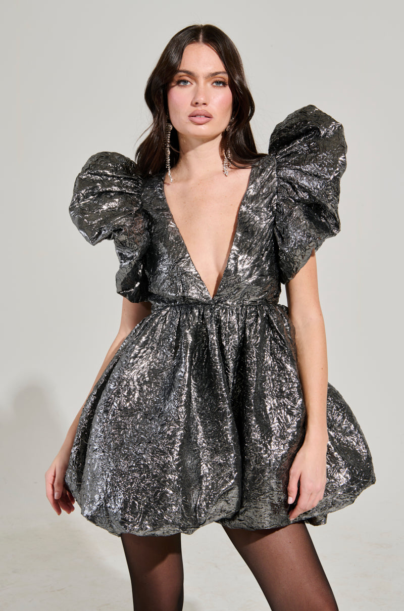 A woman models the BELLE OF THE BALL METALLIC MINI DRESS, showcasing its shiny, metallic fabric and eye-catching puffy sleeves along with a plunging V-neckline. The structured bubble skirt adds a touch of elegance to the short dress. She accessorizes with long, dangling earrings, her long dark hair flowing freely as she stands against a plain background.