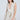 A woman wears the JUST A GIRL TWEED MINI DRESS IN WHITE with a sleeveless, plunging V-neckline and textured plaid. She poses against a plain background, holding a white handbag and wearing large hoop earrings with her long hair complementing the flared skater skirt.