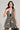 A woman poses confidently in the JUST A GIRL TWEED MINI DRESS, a sleeveless skater design featuring a black and white checkered pattern and a deep V neckline. With long dark hair, she accessorizes with hoop earrings and a small black handbag against a neutral gray background.