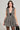 A woman stands wearing the JUST A GIRL TWEED MINI DRESS, which boasts a plaid pattern in black, white, and gold tones with a sequin accent trim and features a deep V-neckline. She holds a black bag and wears hoop earrings, her long wavy hair cascading down. The background is neutral.