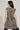 A woman with long, dark hair is shown from behind wearing the JUST A GIRL TWEED MINI DRESS, which features a black, white, and gold plaid sleeveless design. She holds a black handbag and wears gold earrings.