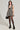 A woman poses in the JUST A GIRL TWEED MINI DRESS, featuring a sleeveless design and deep V-neck with a houndstooth print accented by sequins. She complements her ensemble with a small black square handbag, black sheer tights, and platform heels, while the neutral background highlights her chic attire.