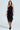 A woman wearing the 90's Baby Velvet Burnout Midi Dress with a purple halter neckline, cinched waist, and fringed hem stands against a plain backdrop. The dress has black textured patterns. She completes the look with black strappy heels, and her long wavy hair flows elegantly.