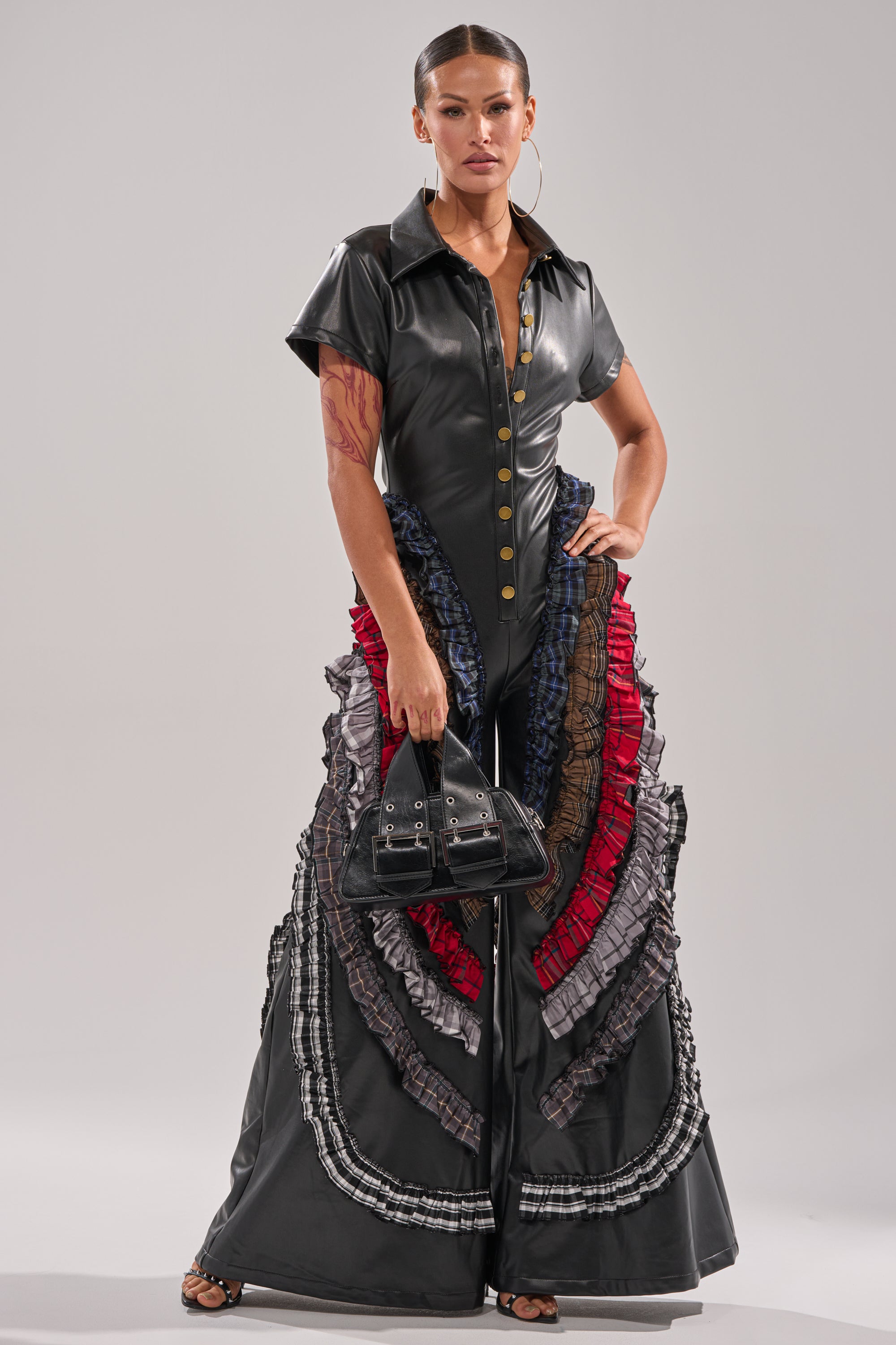 A woman stands confidently in the MAIN CHARACTER ENERGY PU JUMPSUIT featuring bold multicolored ruffle leg accents. She accessorizes with a small black handbag, hoop earrings, and has slicked-back hair with a neutral expression. 0
