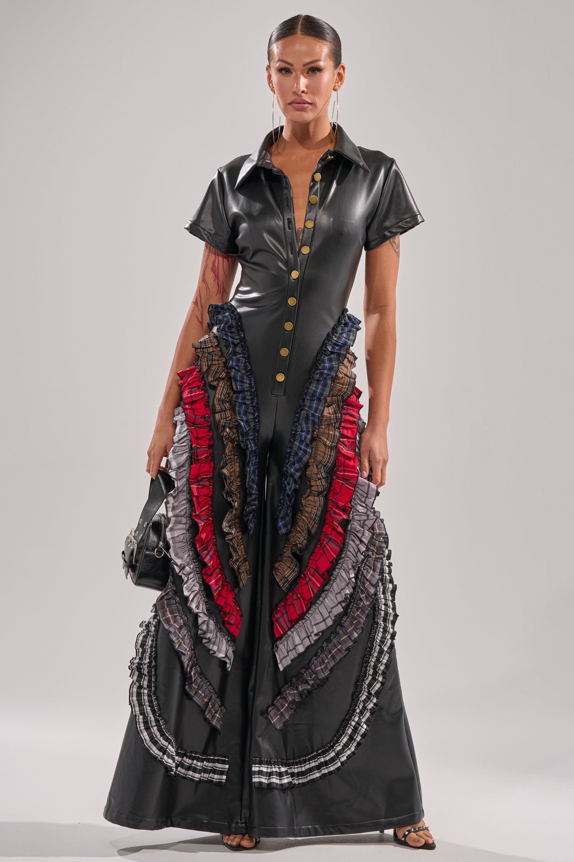 A woman stands facing forward in the MAIN CHARACTER ENERGY PU JUMPSUIT, featuring short sleeves, gold buttons, and colorful ruffled panels in red, purple, blue, and gray cascading down wide legs. She holds a small black handbag. 1