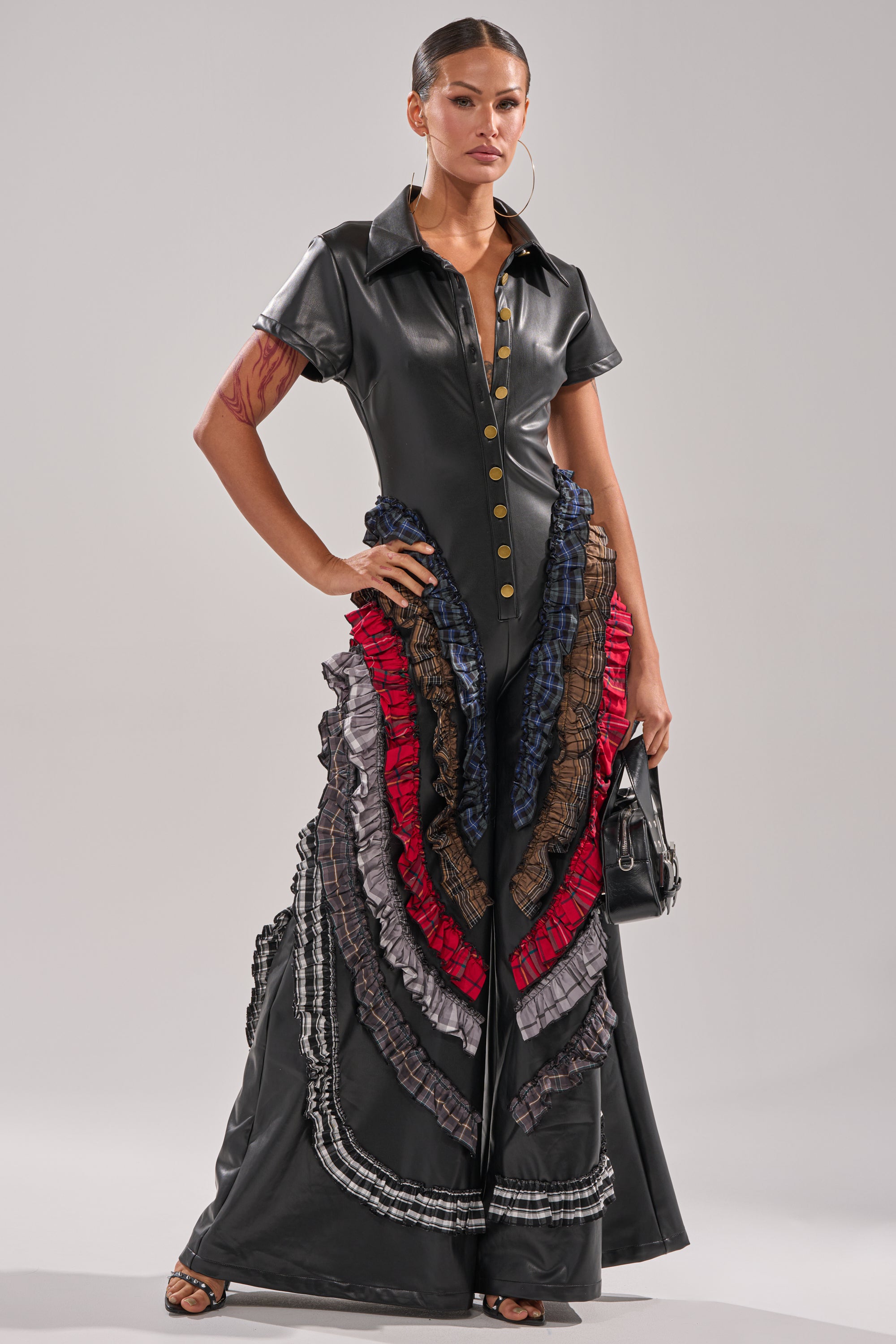 A woman exudes confidence in the MAIN CHARACTER ENERGY PU JUMPSUIT, featuring short sleeves, gold buttons, and layered ruffled accents in red, blue, brown, and gray. She carries a small black handbag. 2