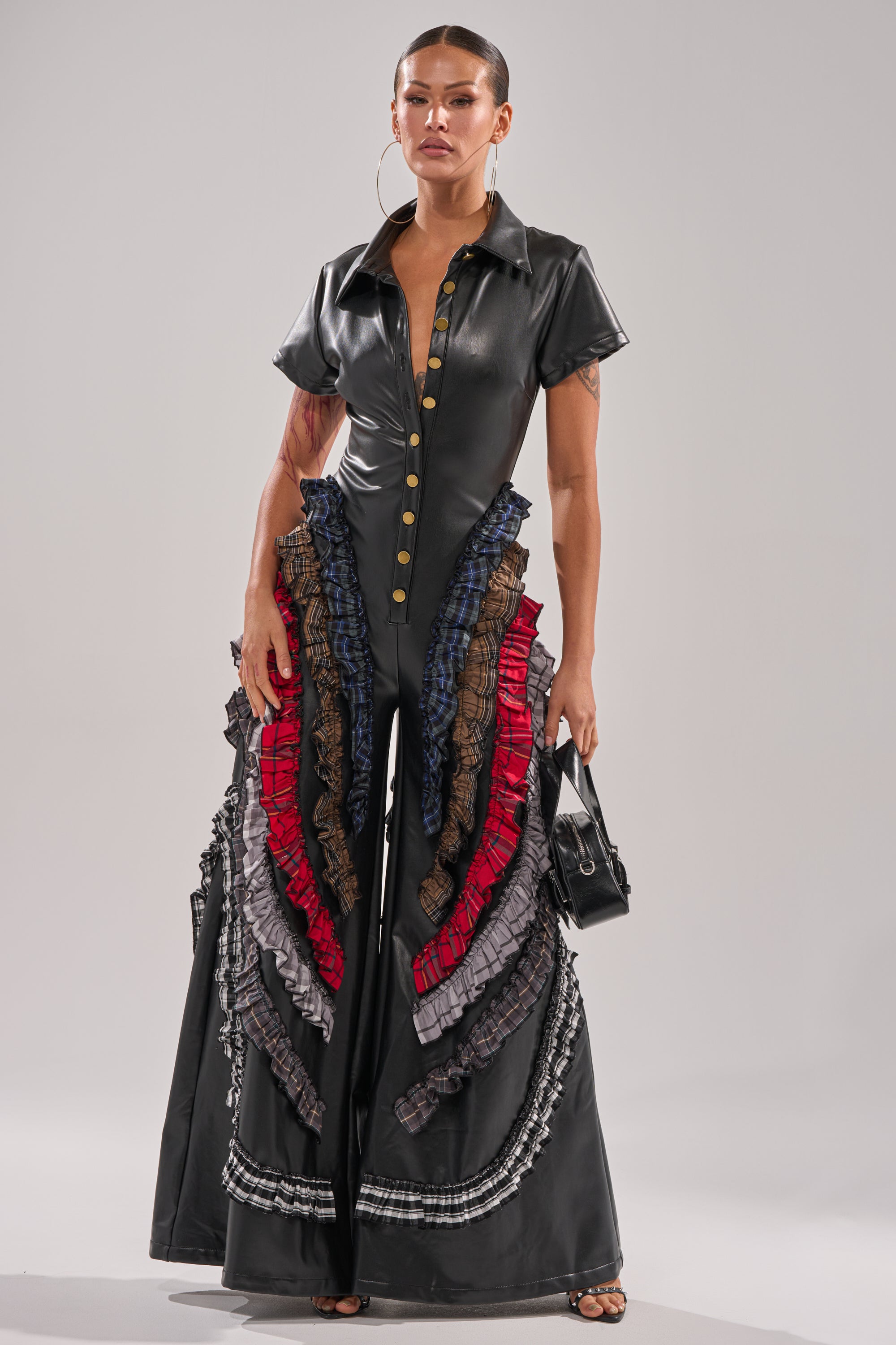 A woman exudes confidence in the MAIN CHARACTER ENERGY PU JUMPSUIT, featuring gold buttons and colorful layered ruffles on wide-leg pants. She accessorizes with a small black handbag, large hoop earrings, and slicked-back hair. 3