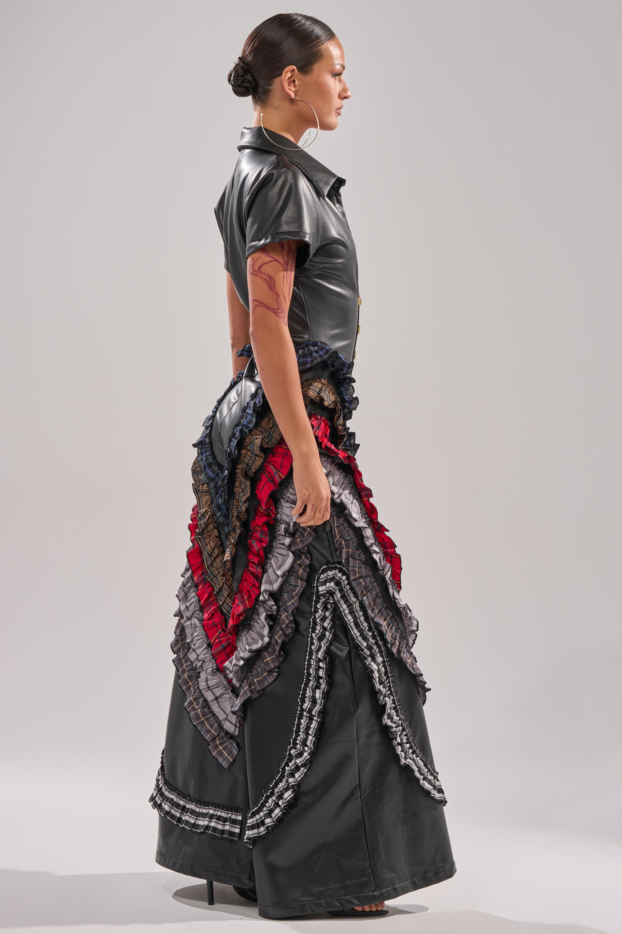 A woman in profile wears the MAIN CHARACTER ENERGY PU JUMPSUIT with short sleeves and a long black skirt adorned with layered, multicolored ruffles. Her hair is pulled back, highlighting a visible arm tattoo. 4