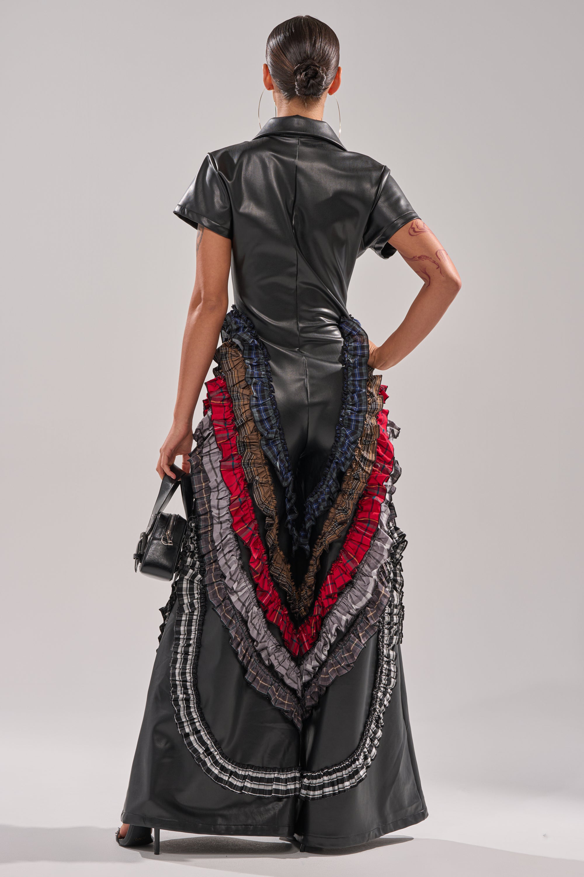 A person faces away from the camera wearing the MAIN CHARACTER ENERGY PU JUMPSUIT, a black faux leather piece with vibrant plaid and solid layered ruffles cascading down the back, accessorized with a small black handbag. 5