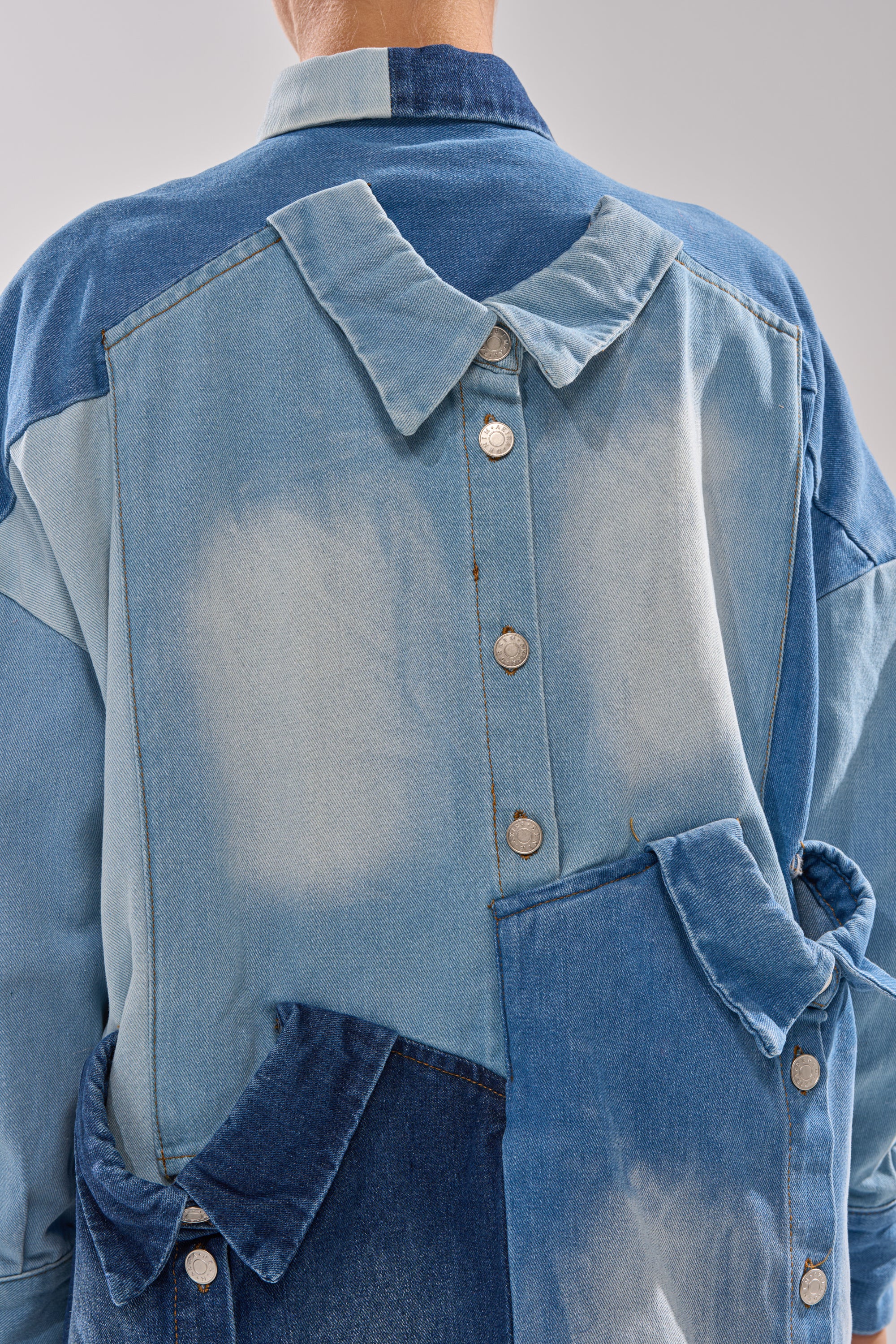 The MONDAY BLUES DENIM MINI DRESS showcases mismatched light and dark blue denim patches, oversized collars, and silver buttons arranged in unique, asymmetric patterns on the back.