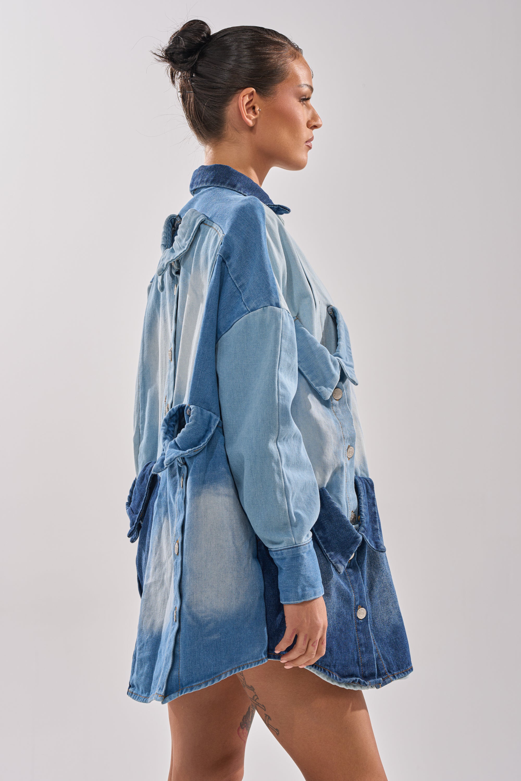 A woman with dark hair in a bun wears the MONDAY BLUES DENIM MINI DRESS, featuring a relaxed fit, patchwork design in various blue shades, large front pockets, and visible seams. She stands sideways against a plain light background.