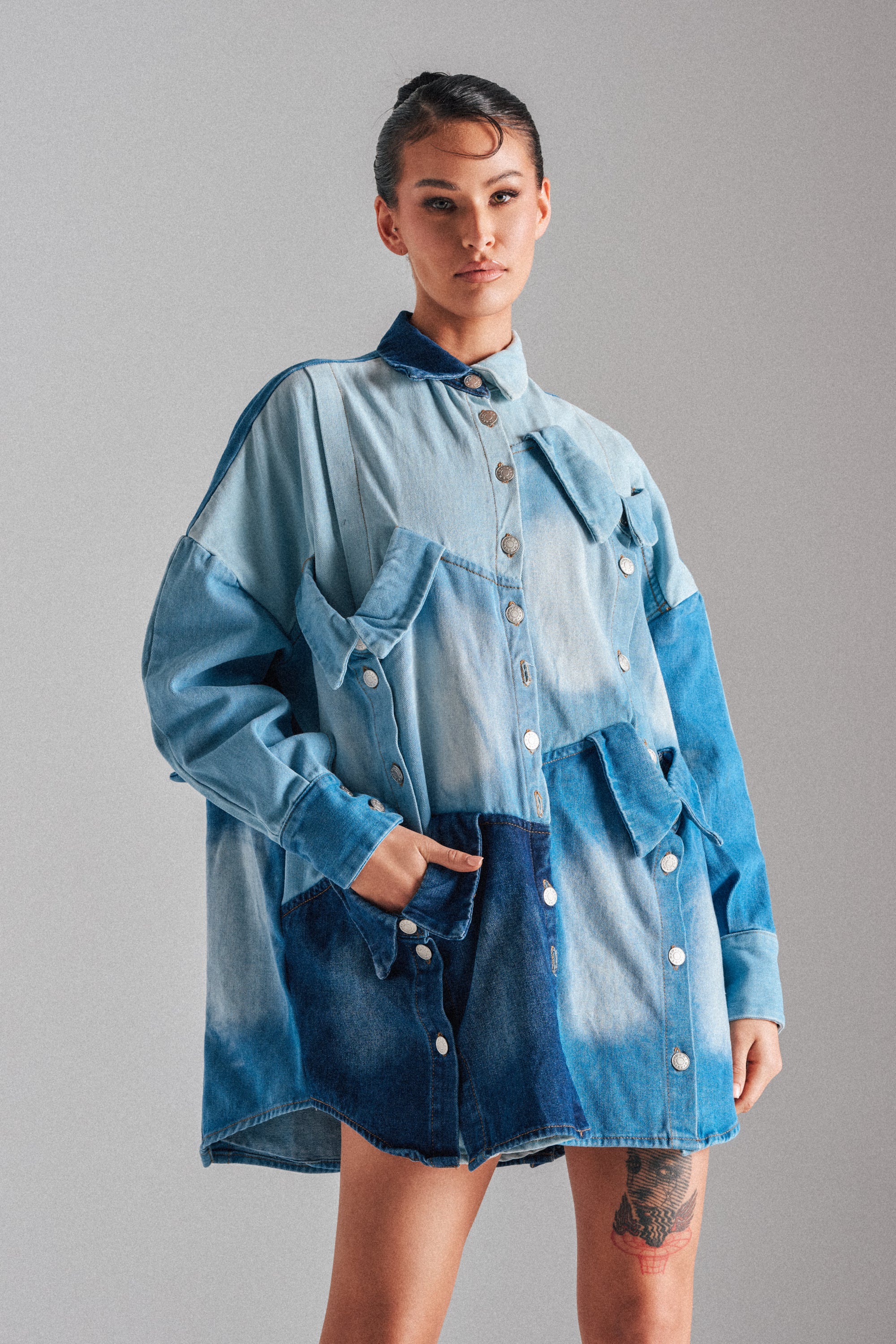 A person stands against a plain background wearing the MONDAY BLUES DENIM MINI DRESS—an oversized patchwork denim dress with large pockets and metal buttons. Their hair is tied back, revealing a thigh tattoo and highlighting the dress’s relaxed silhouette.