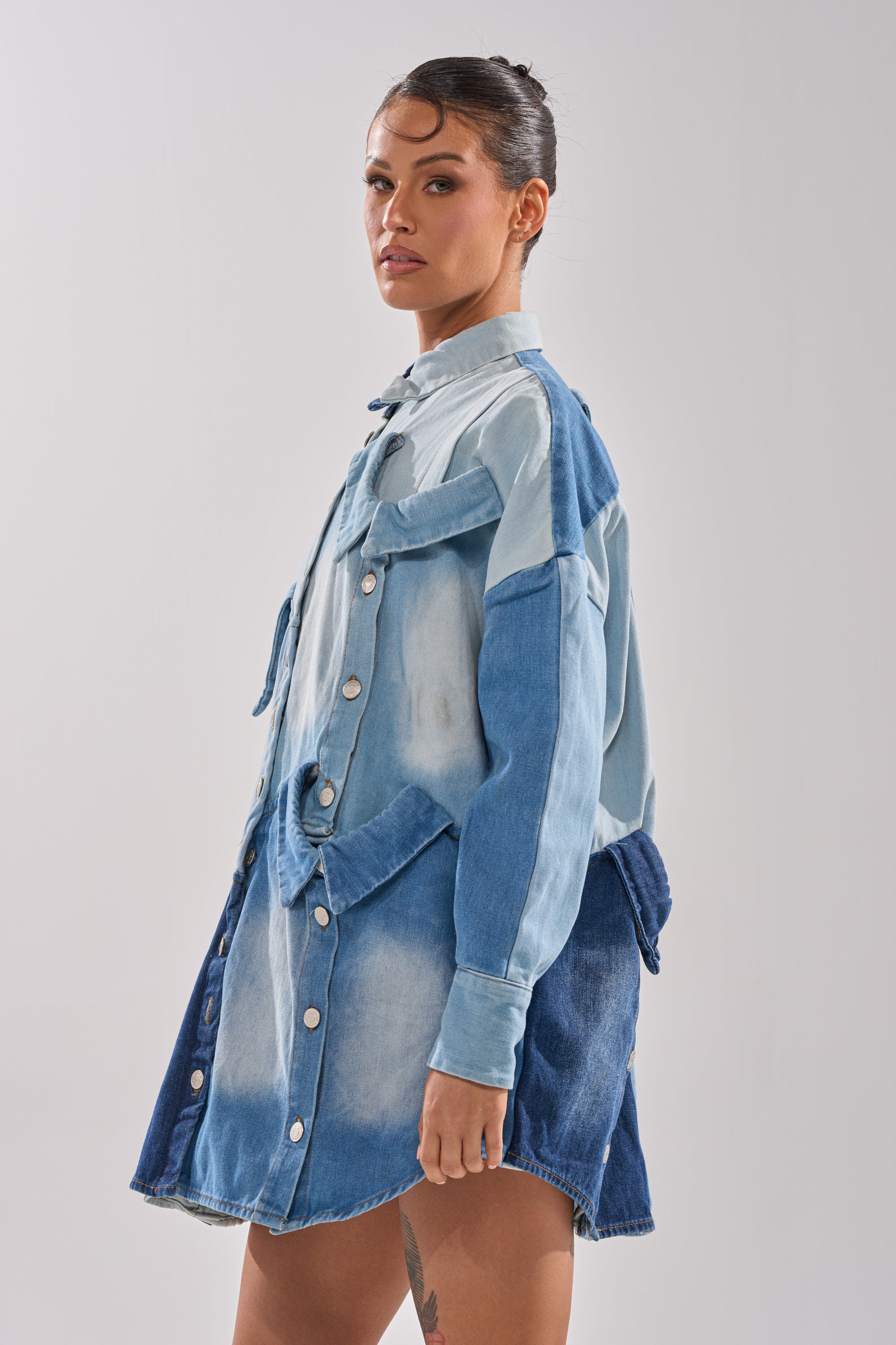 A woman with dark hair pulled back poses confidently in the MONDAY BLUES DENIM MINI DRESS, featuring a relaxed silhouette, long sleeves, snap buttons, and light and dark blue patchwork panels against a plain background.