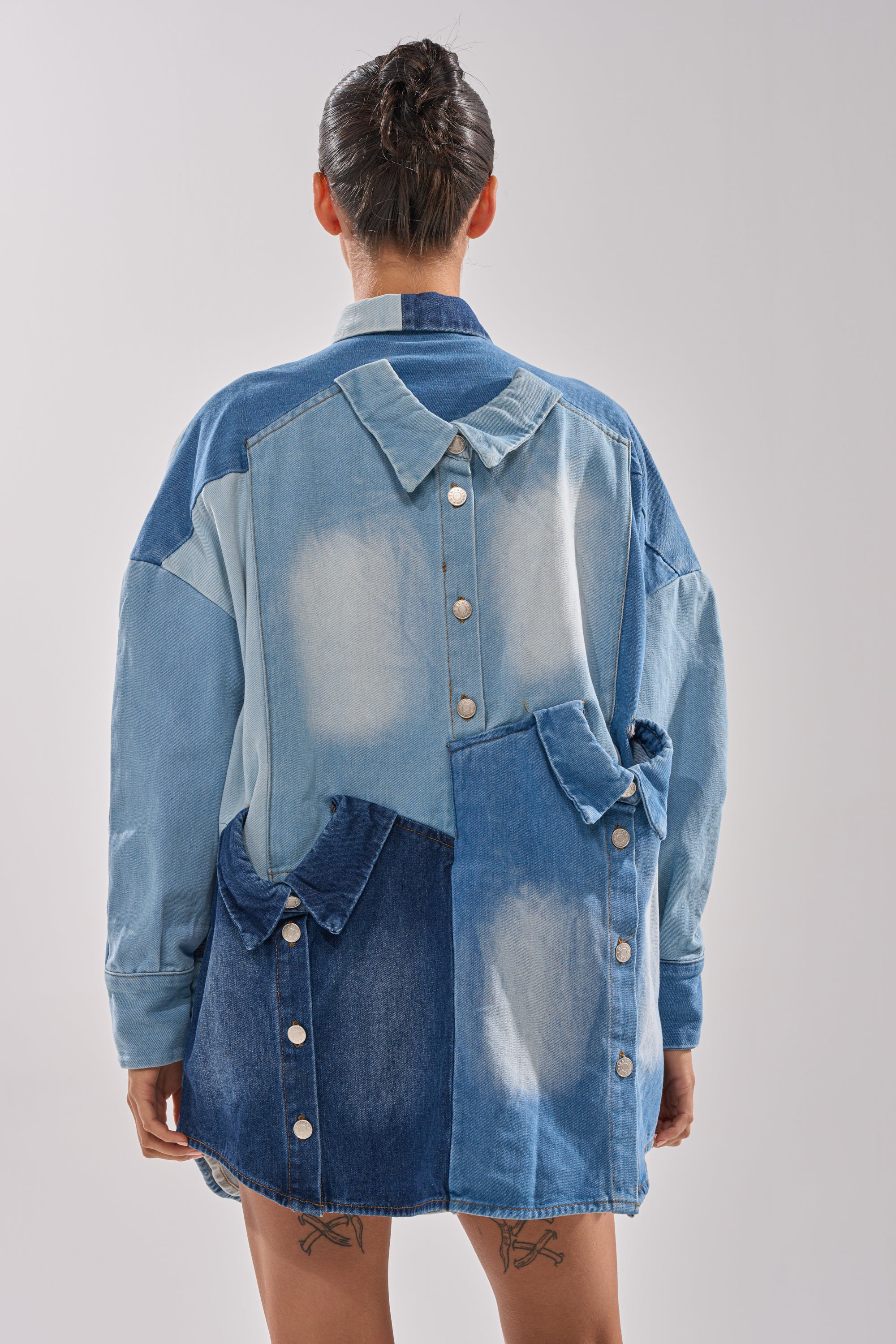 A person with a bun hairstyle is seen from behind wearing the MONDAY BLUES DENIM MINI DRESS, featuring patchwork in various blue shades, buttons, a unique collar and pockets. Tattoos are visible on the backs of their thighs.