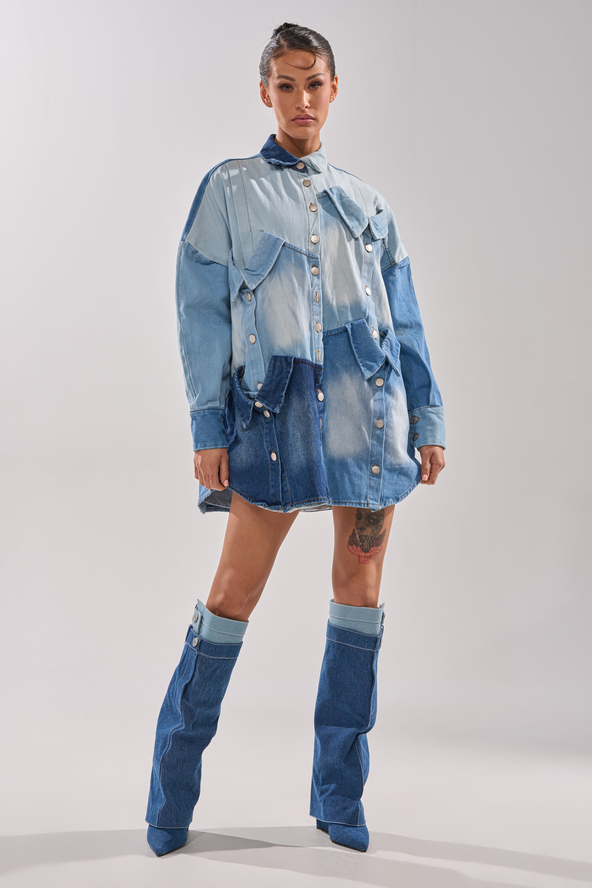 A woman stands confidently in the MONDAY BLUES DENIM MINI DRESS, featuring a relaxed silhouette and color-blocked patchwork design, paired with matching denim boots. The plain background highlights her fashion-forward style.