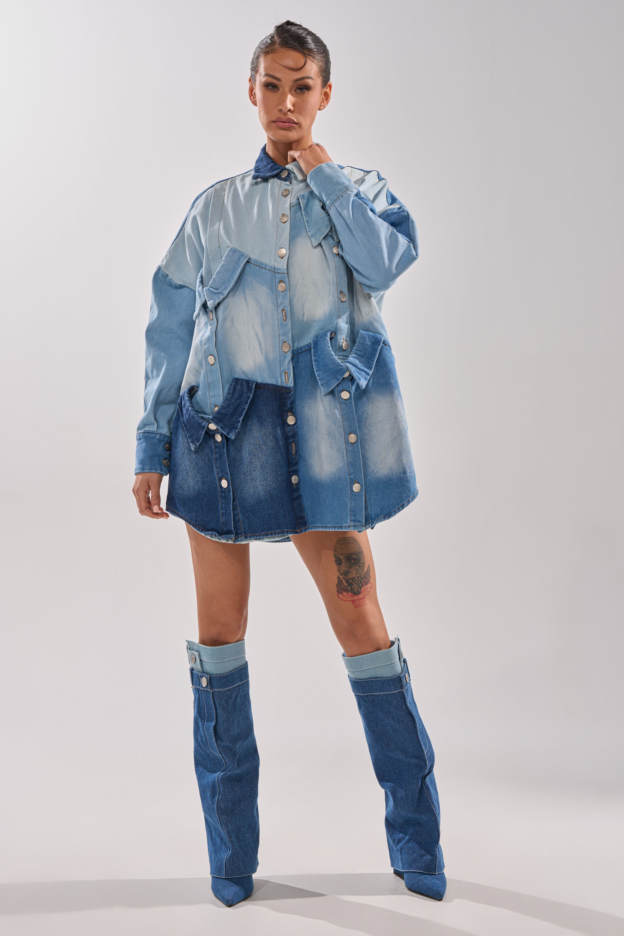 A woman with her hair styled up and a thigh tattoo poses confidently in the MONDAY BLUES DENIM MINI DRESS, paired with tall pointed denim boots, against a plain light background.