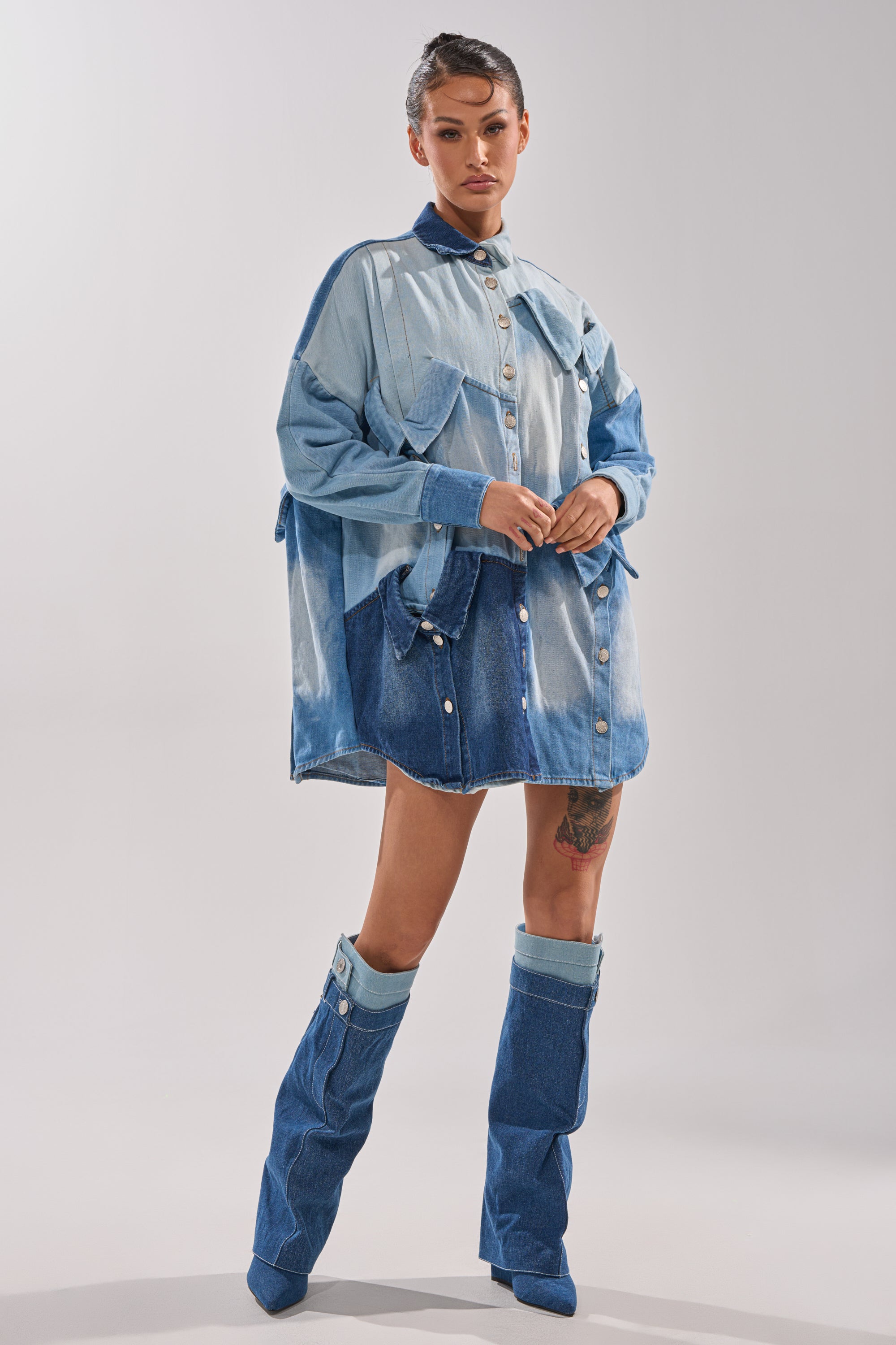 A woman poses confidently in the MONDAY BLUES DENIM MINI DRESS, featuring an oversized patchwork design and large pockets, paired with matching knee-high denim boots; she stands against a plain white background.