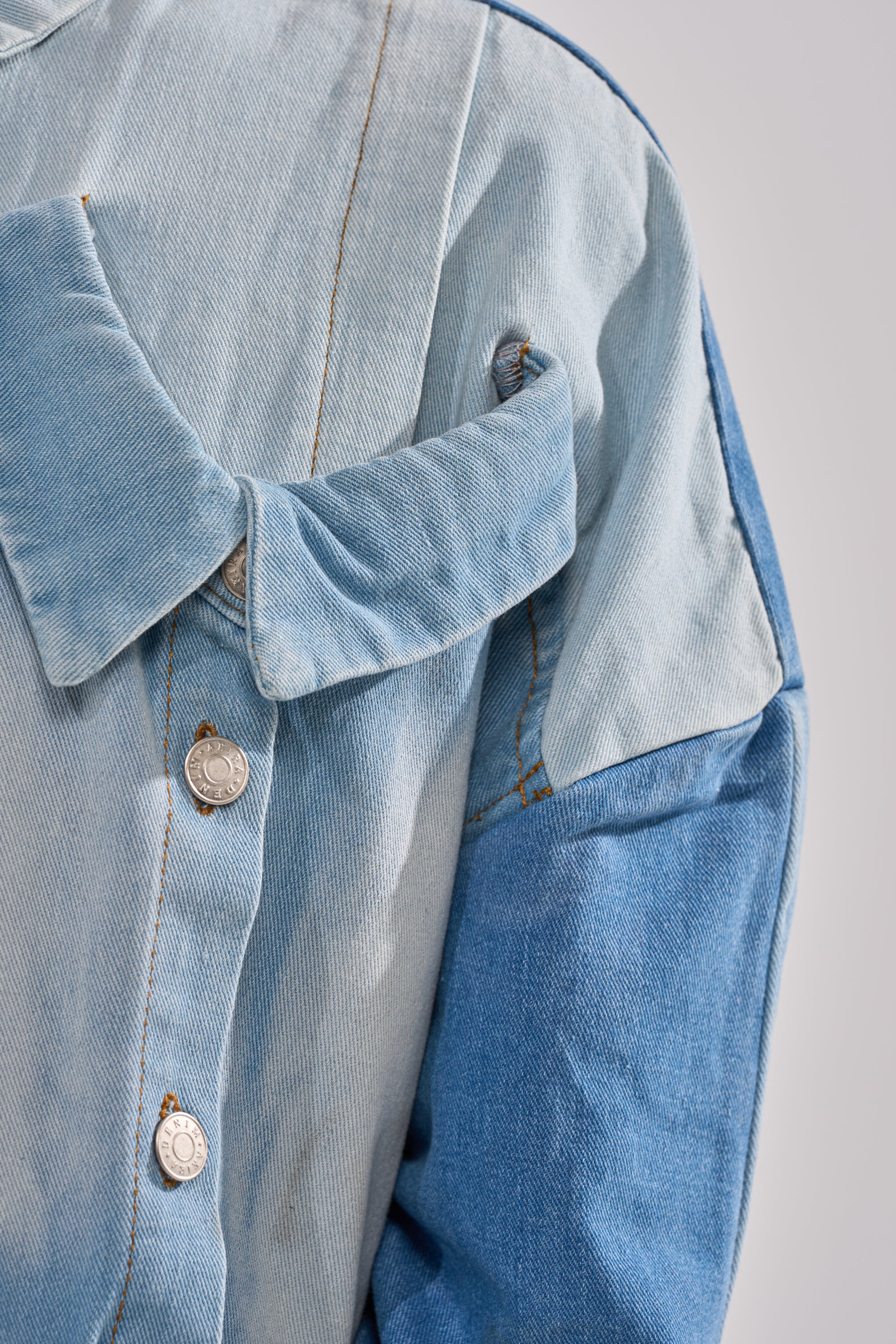 A close-up of the MONDAY BLUES DENIM MINI DRESS, featuring light and dark blue patchwork, metallic buttons, an oversized collar, and visible stitching against a light grey background.