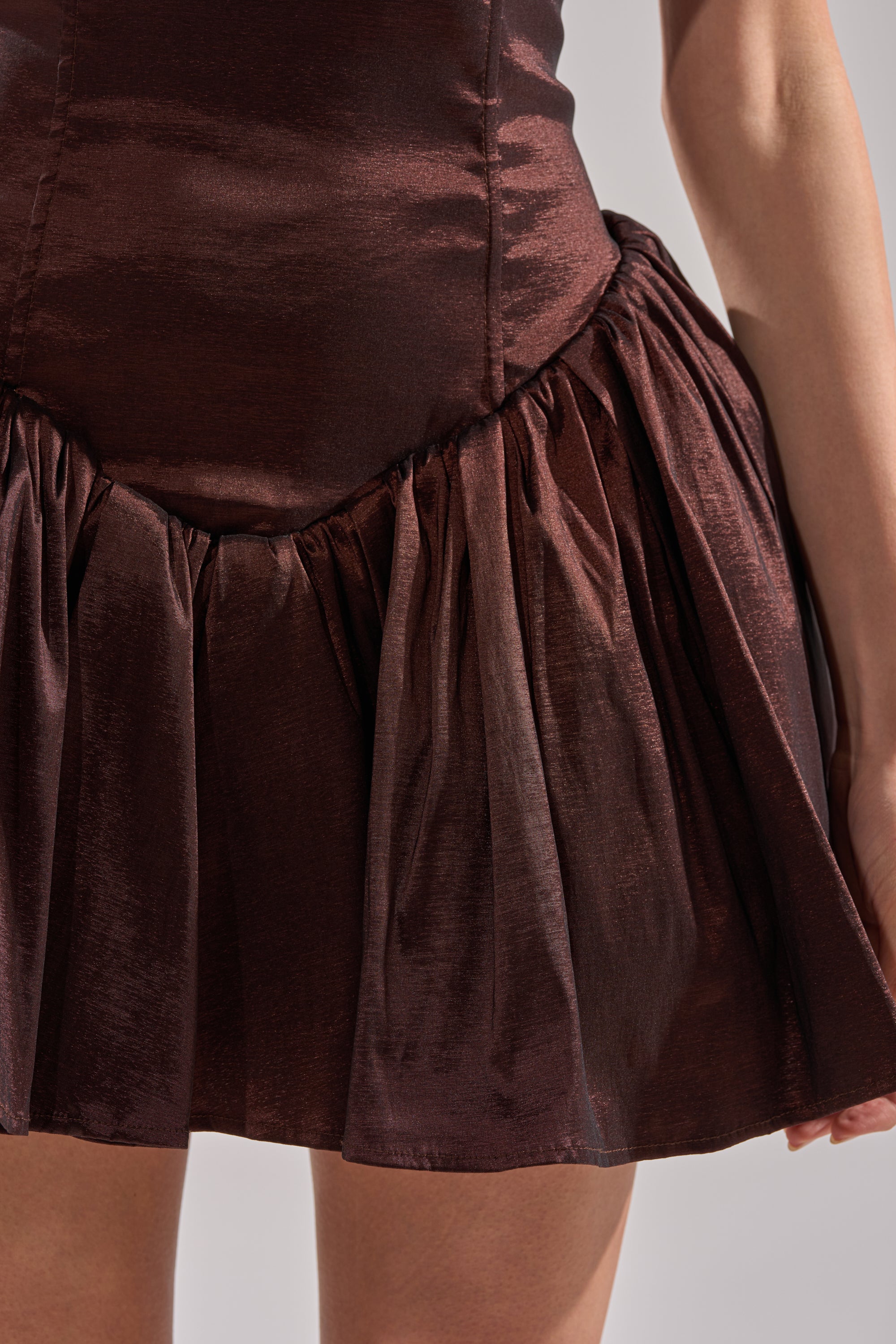 A close-up of a person wearing the I LIKE IT LIKE THAT MINI DRESS, featuring a shiny dark brown fabric, fitted bodice, and gathered flared bubble skirt. Only the torso, upper legs, and part of one arm are visible.