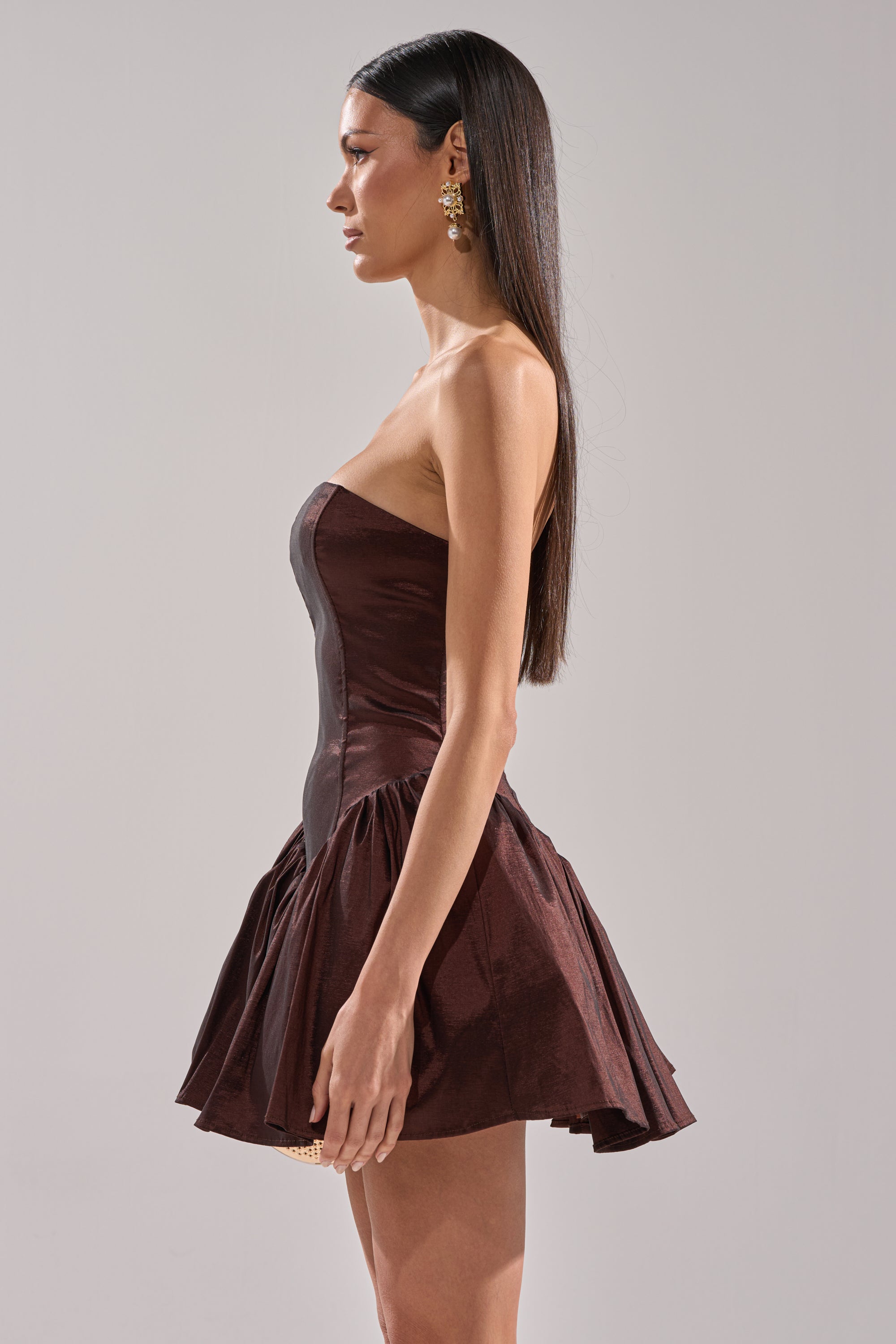 A woman with long straight hair stands in profile wearing the I LIKE IT LIKE THAT MINI DRESS, a strapless brown mini with a flared bubble skirt and gold statement earrings, against a plain light background.
