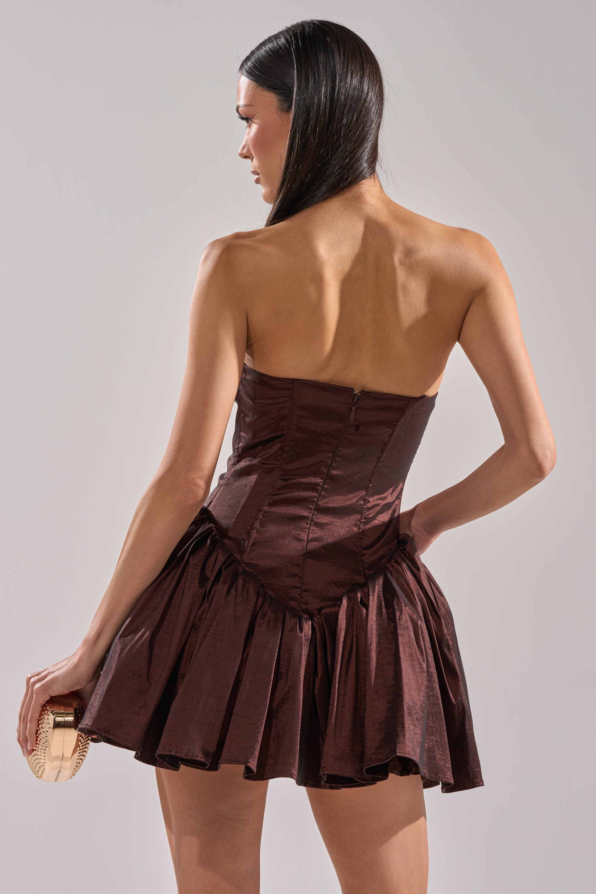 A woman with long dark hair models the I LIKE IT LIKE THAT MINI DRESS, a strapless brown mini dress with a flared bubble skirt. She stands with her back to the camera, holding a light-colored clutch in her right hand.