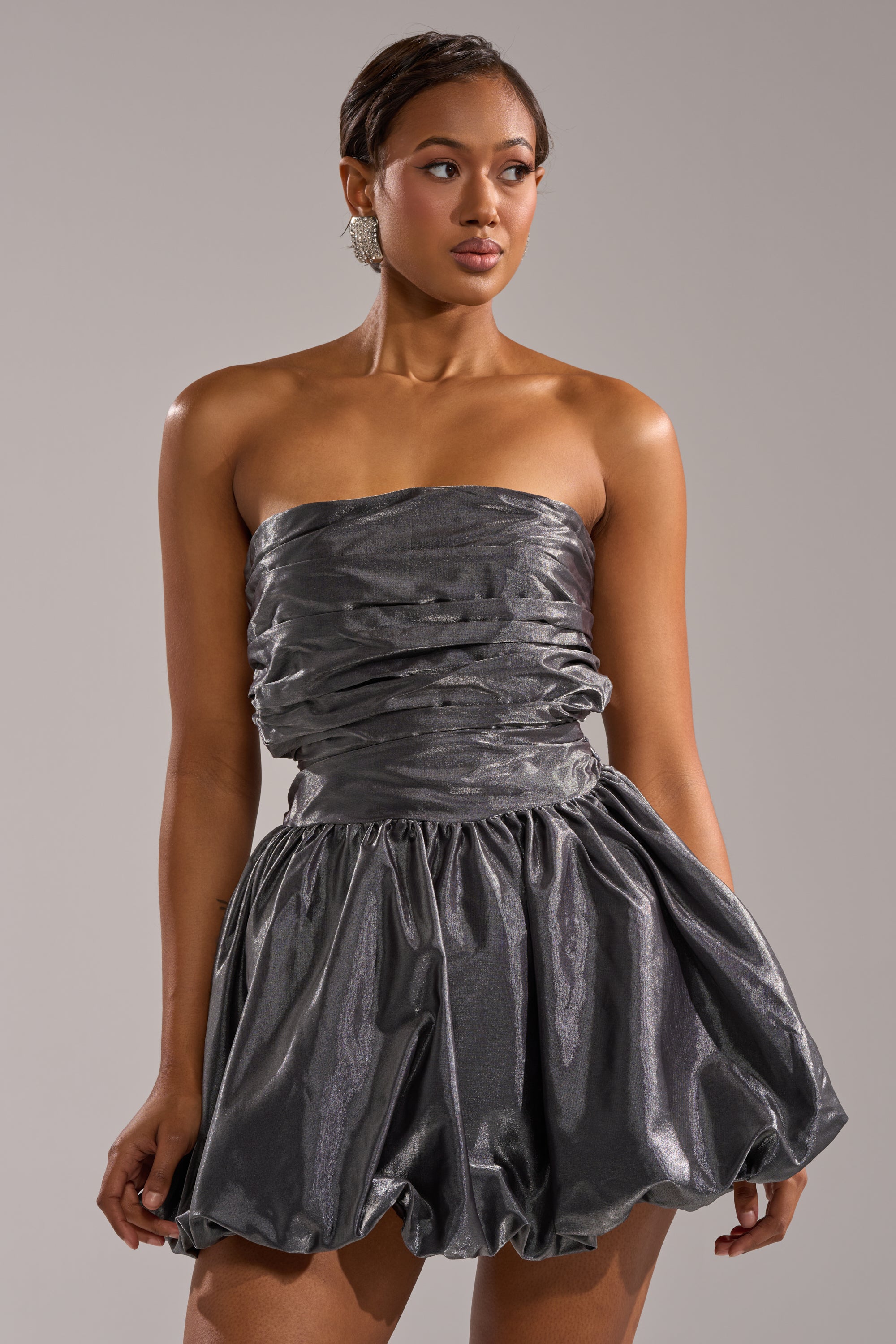 A woman models the GET WITH IT MINI DRESS, featuring a strapless metallic design with a ruched bodice and voluminous bubble skirt, as she stands against a plain light gray background looking slightly to her left. 0