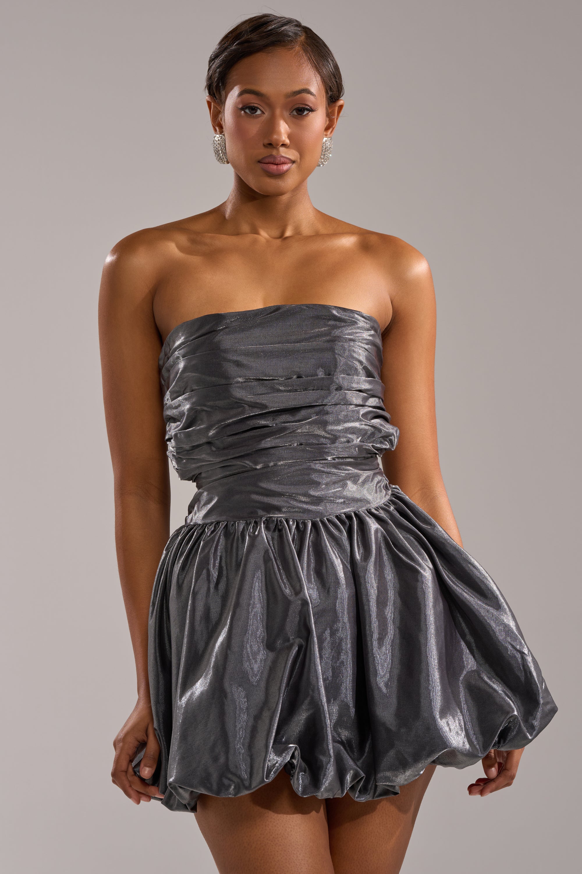 A woman wears the GET WITH IT MINI DRESS—a shiny, strapless metallic mini with a ruched bodice and voluminous bubble skirt—standing against a plain light background; an eye-catching choice for parties and celebrations. 1