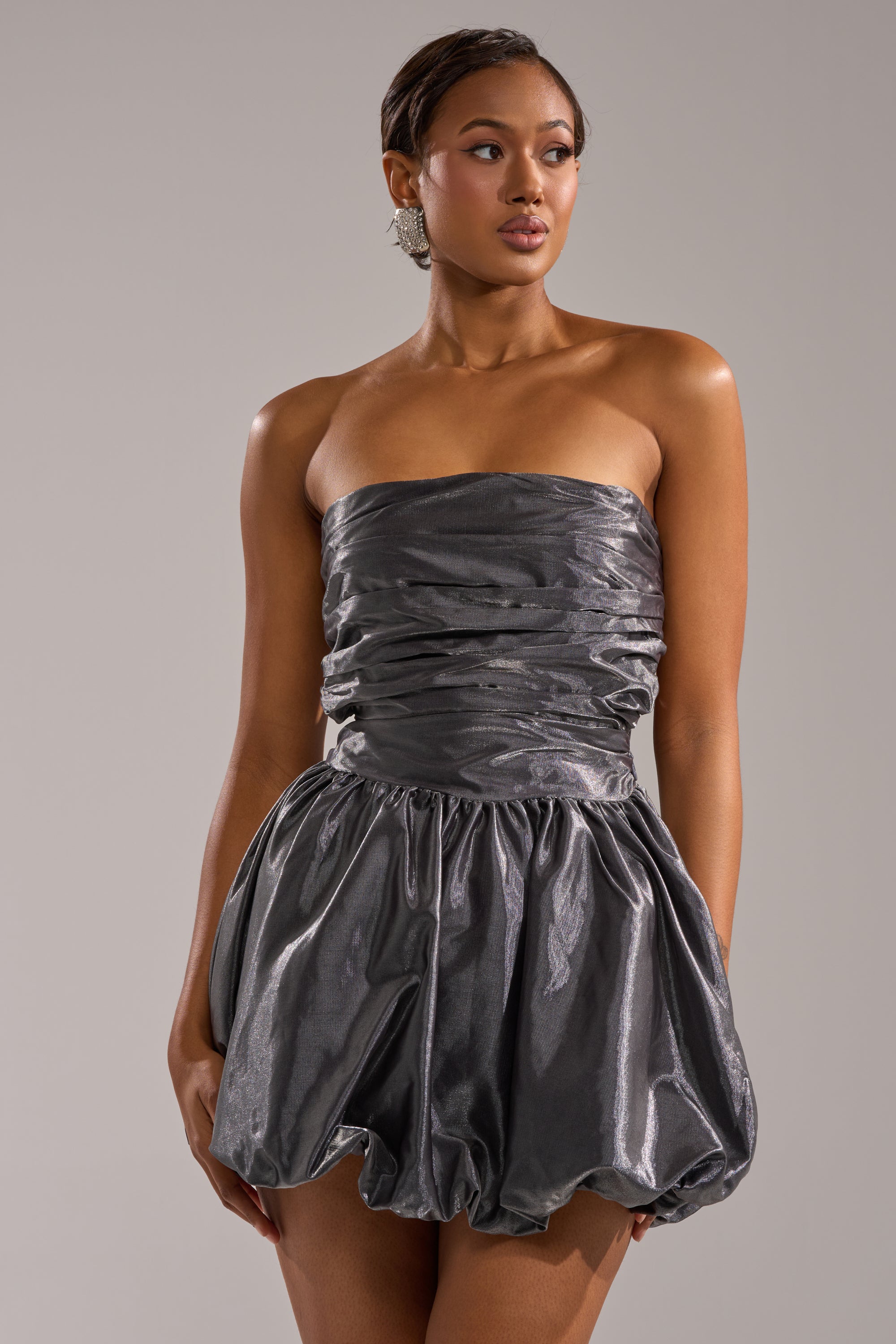 A woman stands against a neutral background, wearing the GET WITH IT MINI DRESS—a strapless metallic mini with a ruched bodice and voluminous bubble skirt—looking slightly to the side in this eye-catching party look. 3