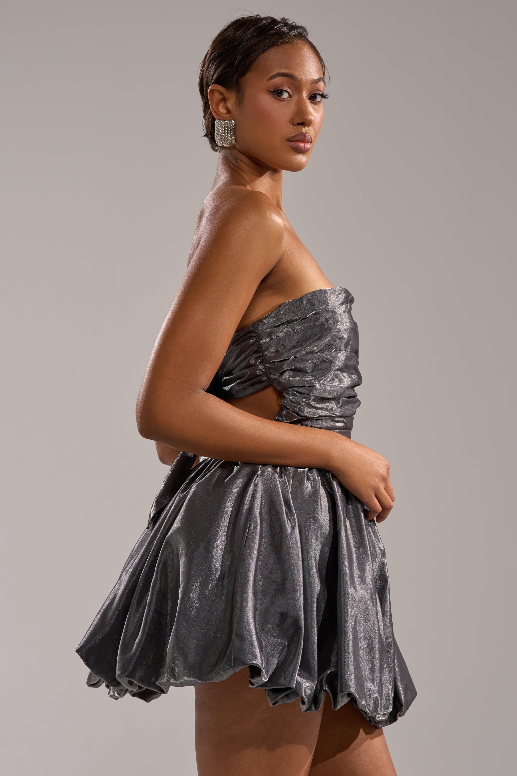 A woman models the GET WITH IT MINI DRESS, a strapless metallic mini, against a light gray backdrop. She accessorizes with bold square earrings and sports neat short hair—serving confident party dress inspiration. 4