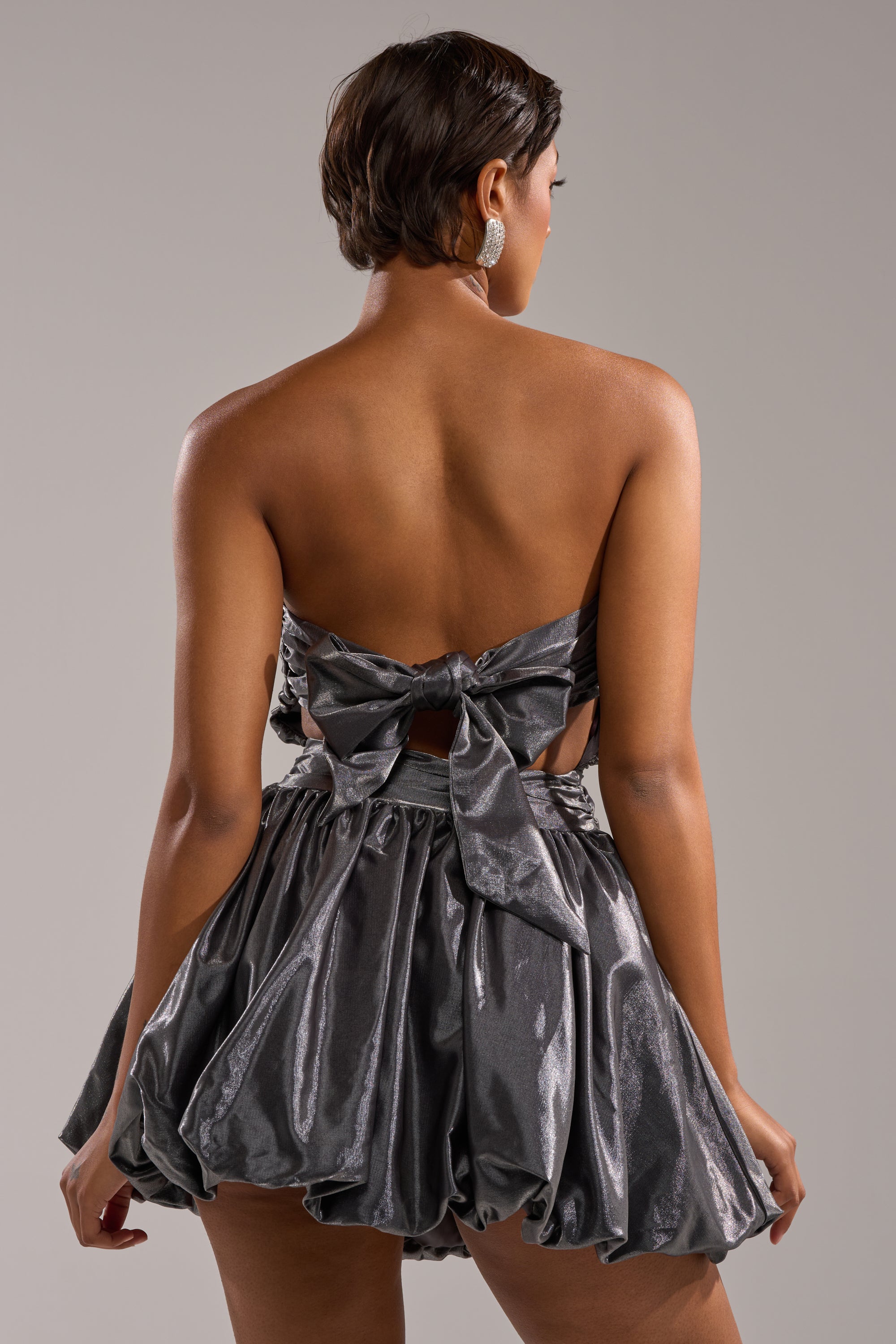 A woman with short hair is seen from the back wearing the GET WITH IT MINI DRESS—a strapless metallic mini dress featuring a large bow and voluminous gathered skirt, making it a standout party look against a neutral backdrop. 5