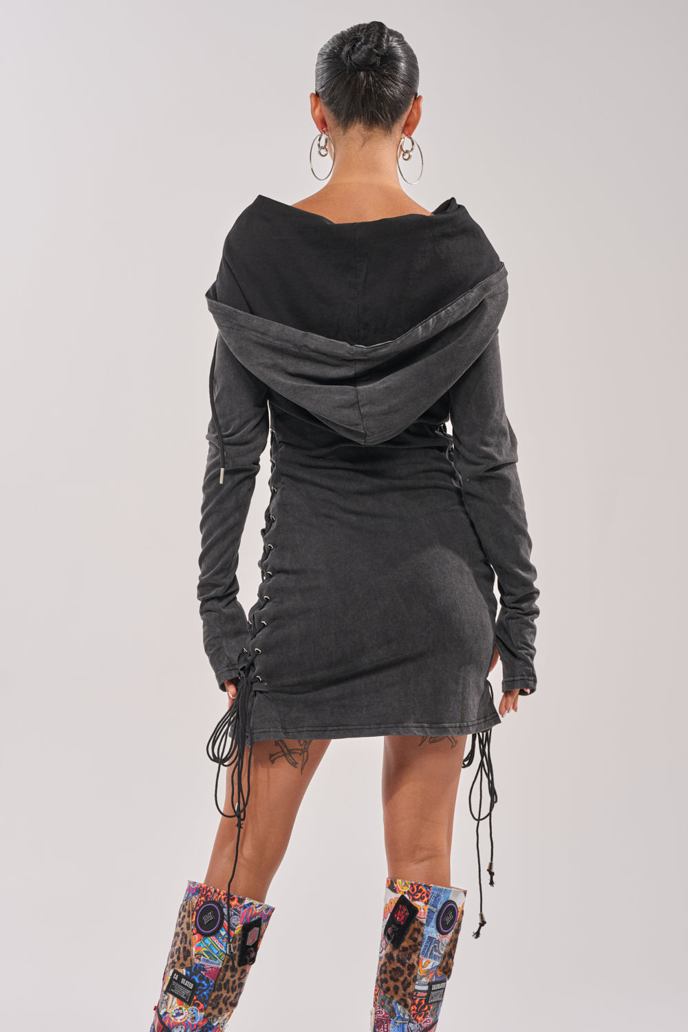 A person with short hair faces away, wearing the BUTTERFLY EFFECT MINI DRESS with side lace-up details and colorful patterned knee-high boots. The background is plain and light-colored. 5