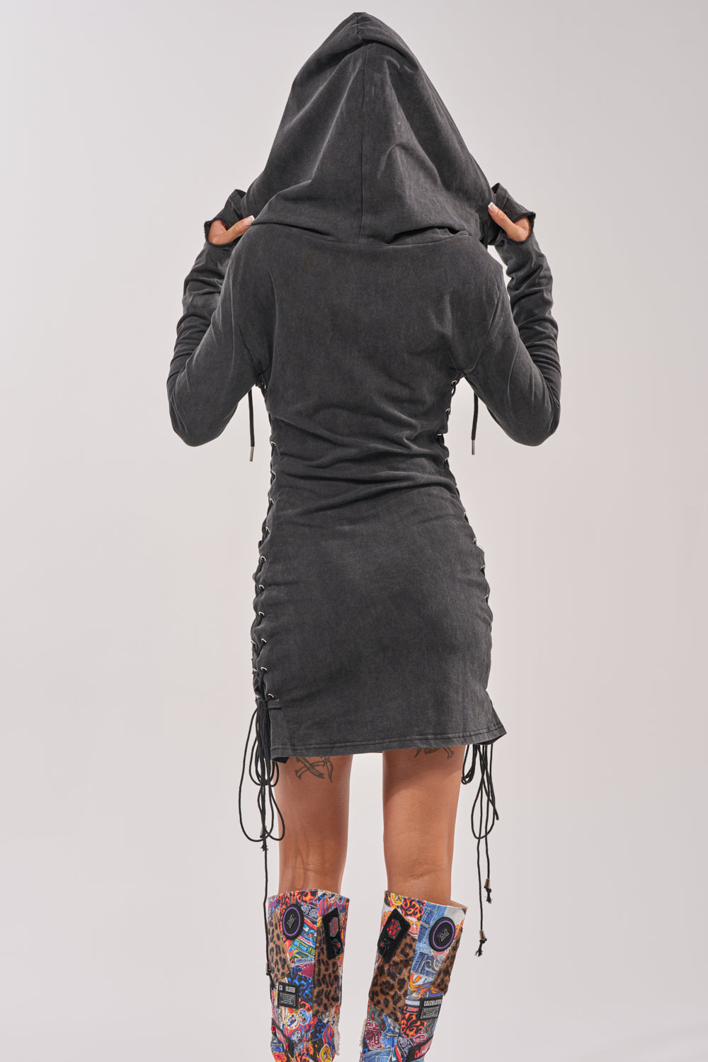 A person faces away, wearing the BUTTERFLY EFFECT MINI DRESS—a ruched black sweatshirt dress with side drawstrings—paired with colorful patterned knee-high boots against a plain light background. 6