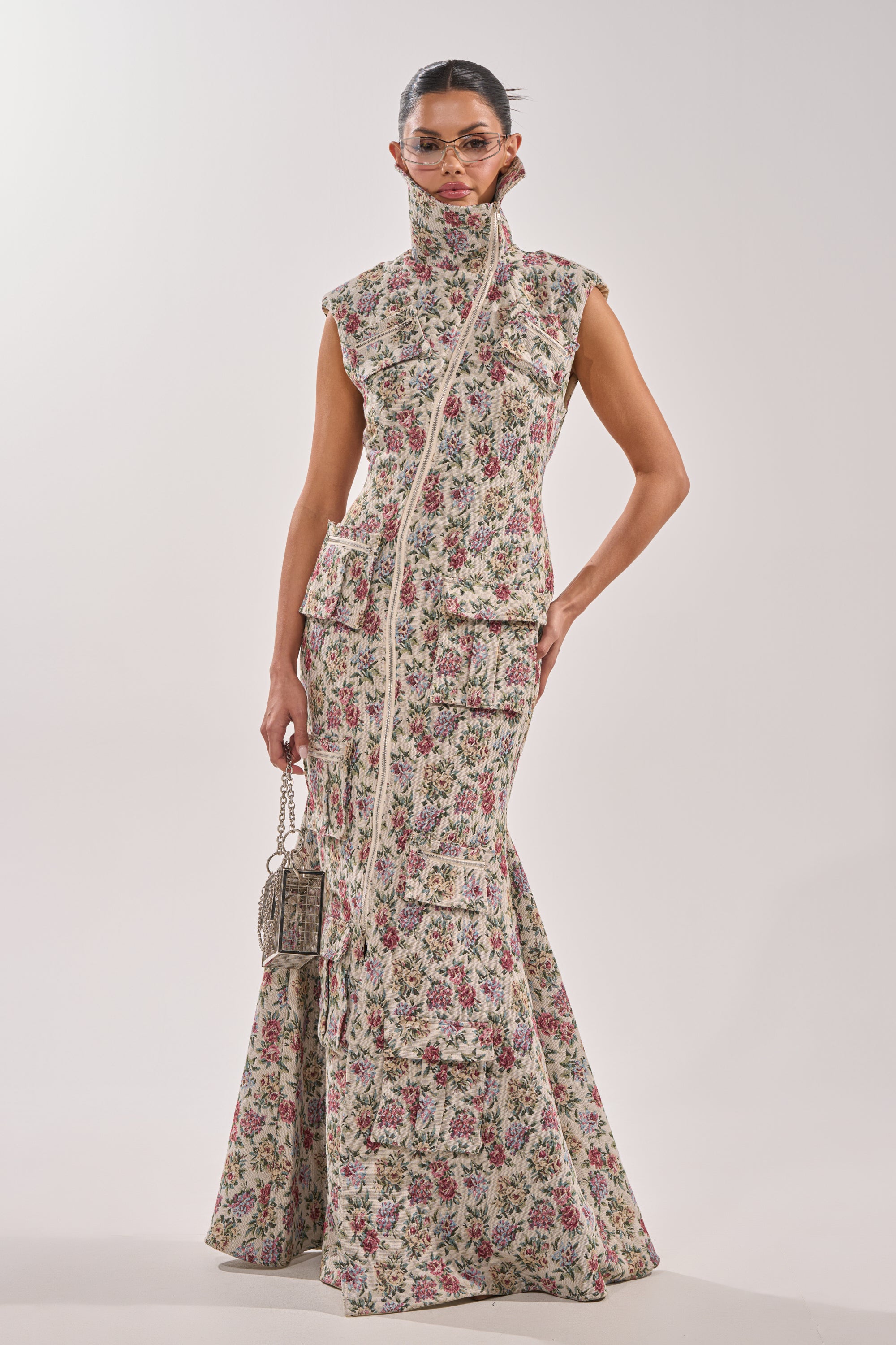 Wearing the FLOWER POWER TAPESTRY MAXI DRESS with a sleeveless design, high asymmetrical zip-up collar, and multiple large pockets, a person holding a small handbag stands against a plain white background. 0