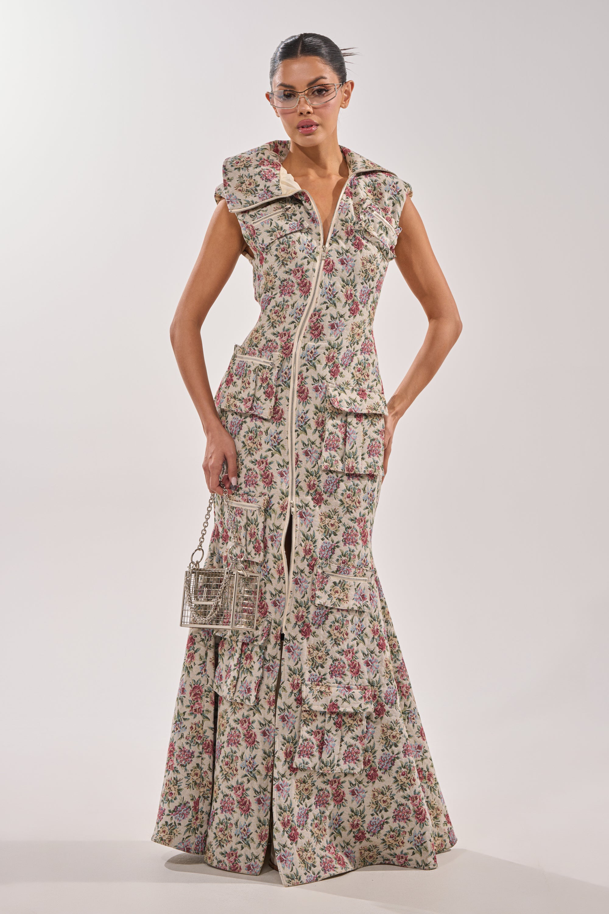 A woman stands against a plain background wearing the FLOWER POWER TAPESTRY MAXI DRESS, a sleeveless floral print style with large front pockets. She pairs it with a small metallic handbag and clear-framed glasses. 2
