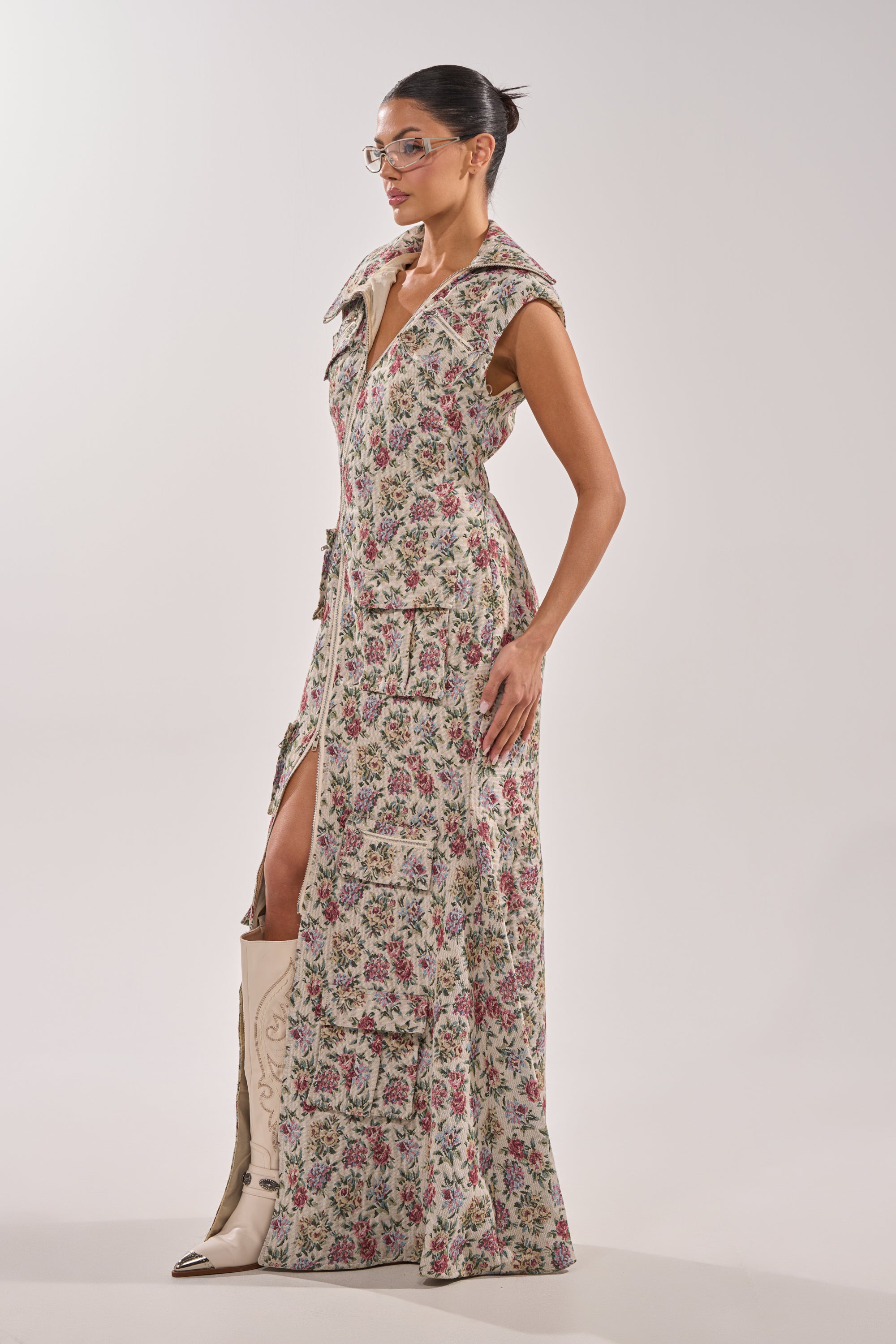 A woman wearing clear glasses and the FLOWER POWER TAPESTRY MAXI DRESS with a large collar and pockets poses confidently, her hair in a neat bun. She completes the look with tall beige boots against a plain light background. 3