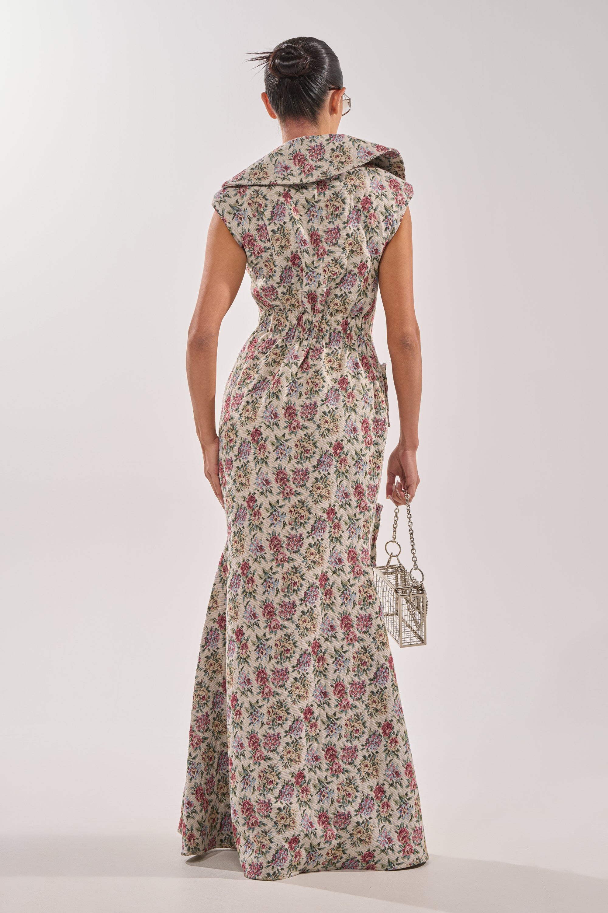 A person with their hair in a bun, facing away, wears the FLOWER POWER TAPESTRY MAXI DRESS—long, sleeveless, with a wide collar and floral pattern. They hold a small boxy handbag against a plain light background. 4