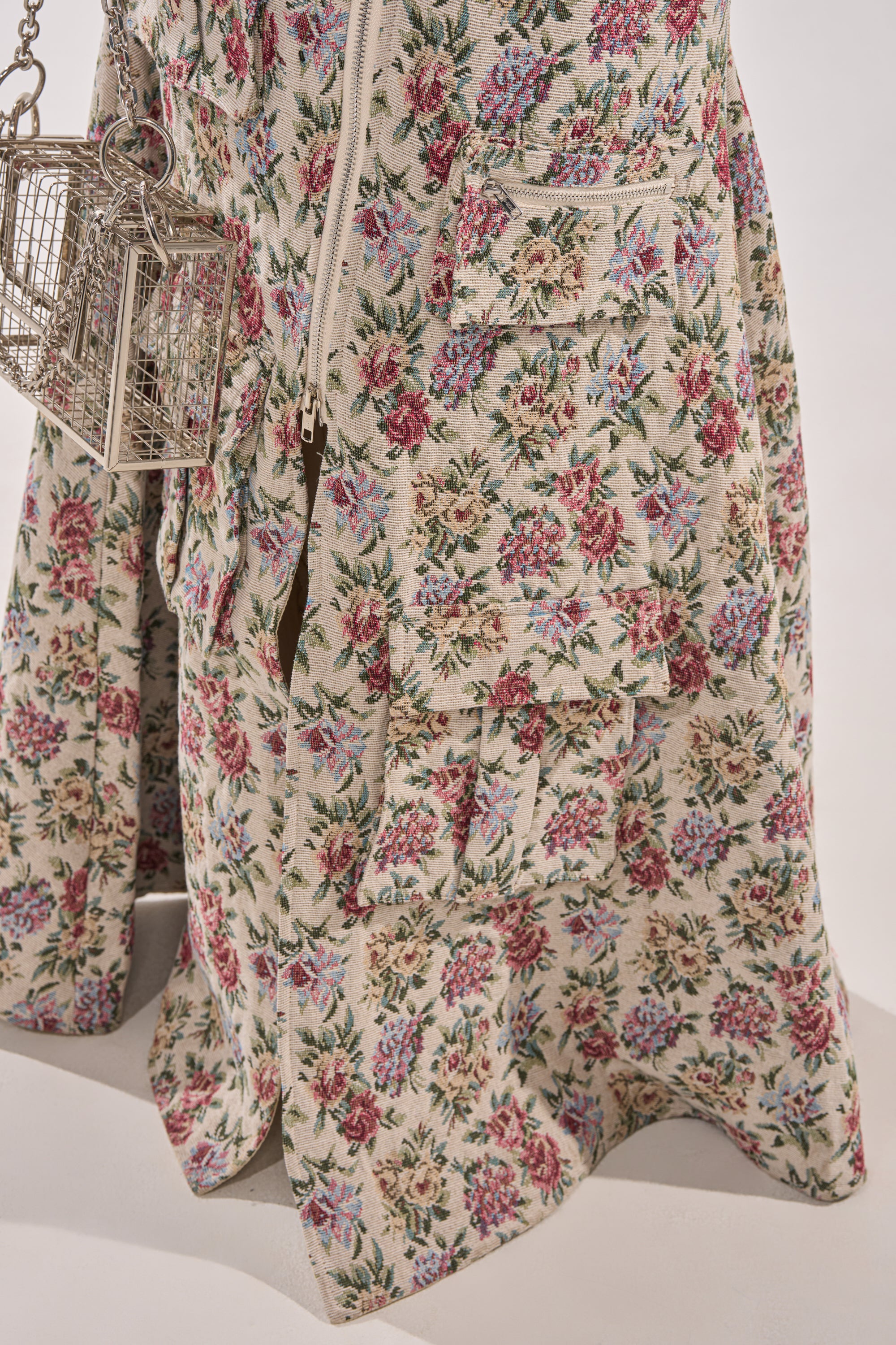 A close-up of the FLOWER POWER TAPESTRY MAXI DRESS, a cream maxi dress with jacquard floral print in pink, red, and blue. Zippered pockets accent the design, while a metallic birdcage-shaped handbag hangs at the side. 6