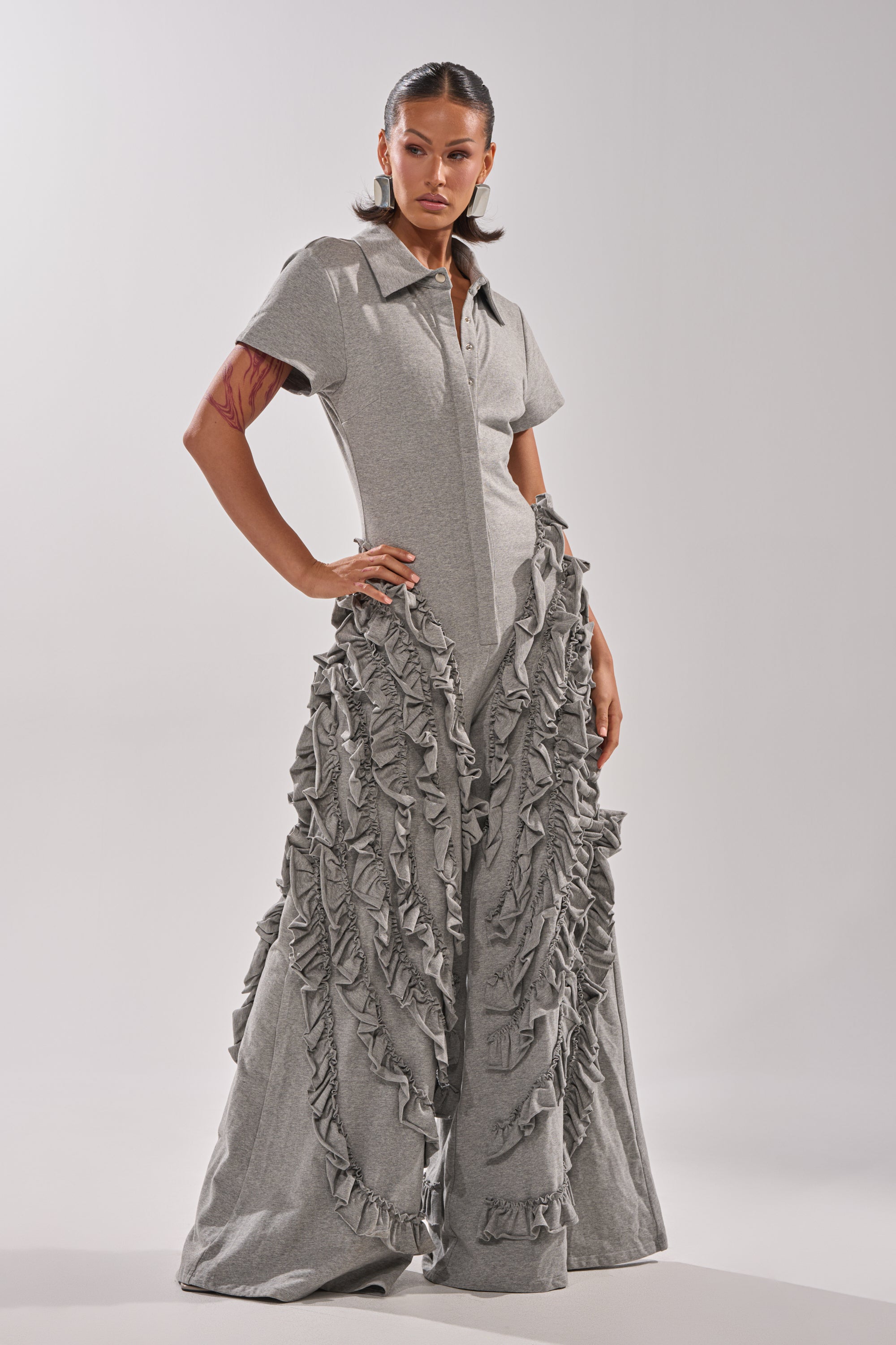 A woman stands confidently in the LEAH RUFFLE JUMPSUIT, a light grey wide-leg jumpsuit featuring a collar, short sleeves, zippered front, and cascading ruffles. She poses against a well-lit plain background.