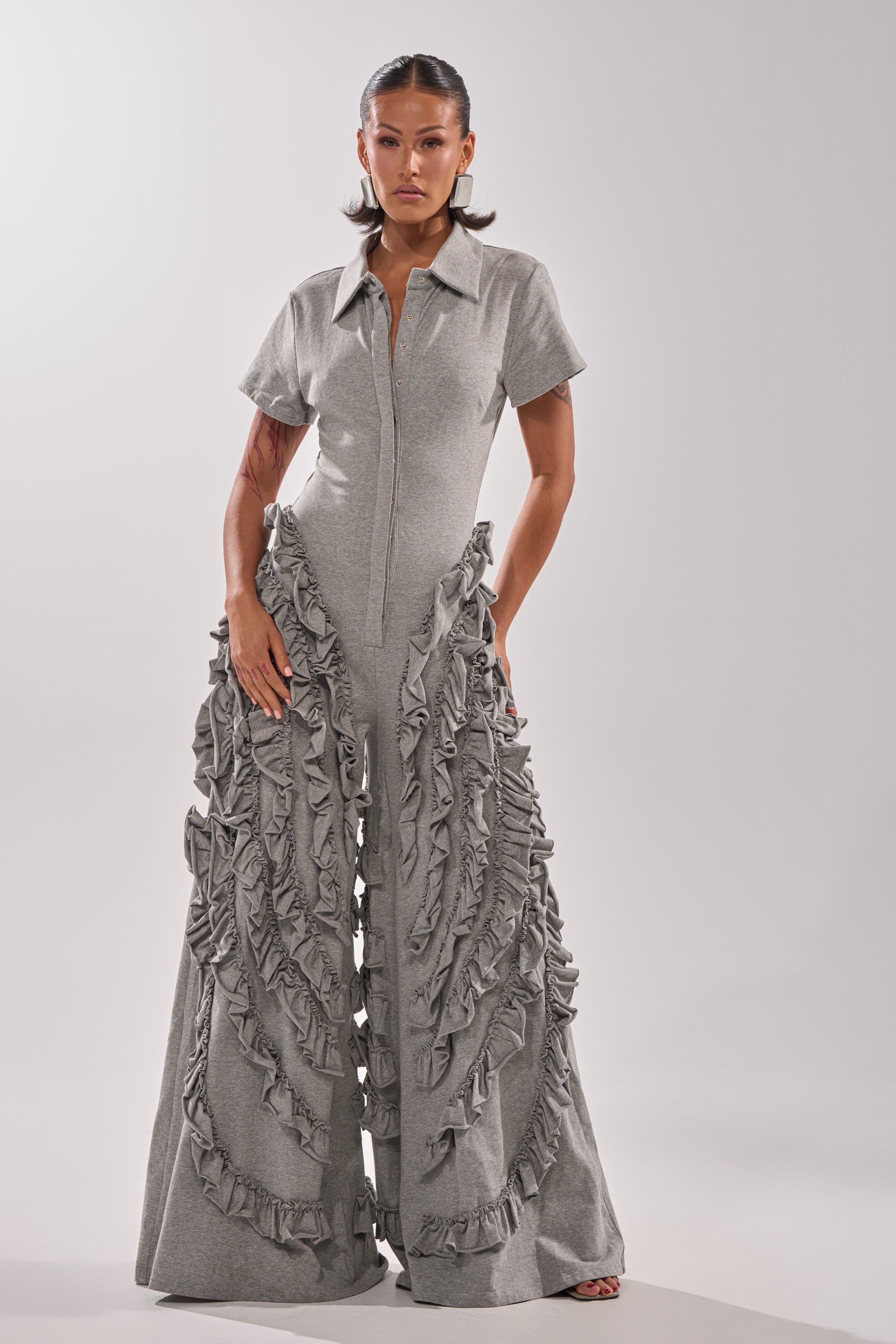 A woman faces forward in the LEAH RUFFLE JUMPSUIT, featuring light gray fabric, wide legs, short sleeves, a collar, and cascading ruffles. The plain white background highlights her standout outfit.