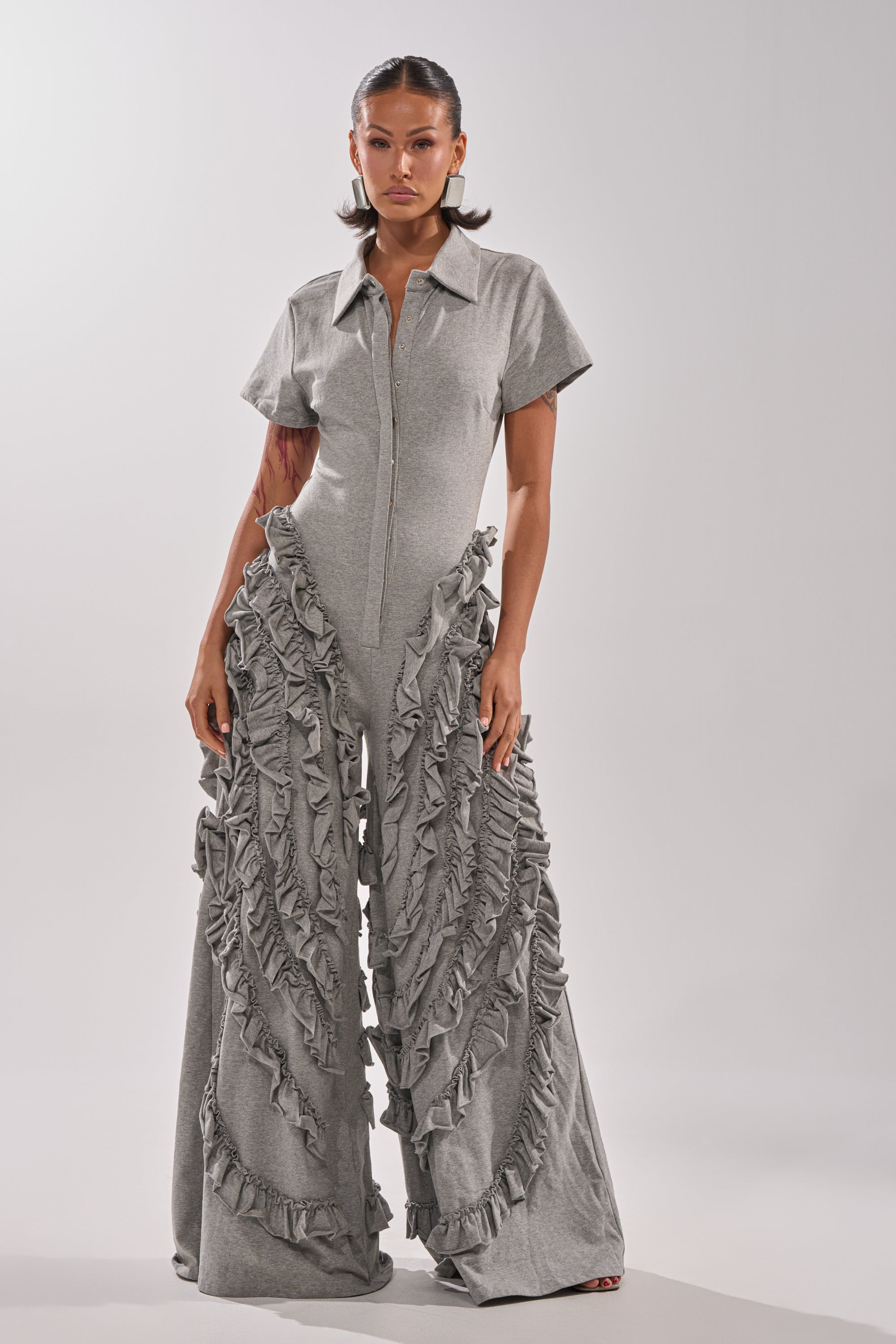 A woman stands confidently in the LEAH RUFFLE JUMPSUIT, a light gray wide-leg piece with short sleeves, a zip-up front, and striking cascading ruffles. The background is plain and neutral.