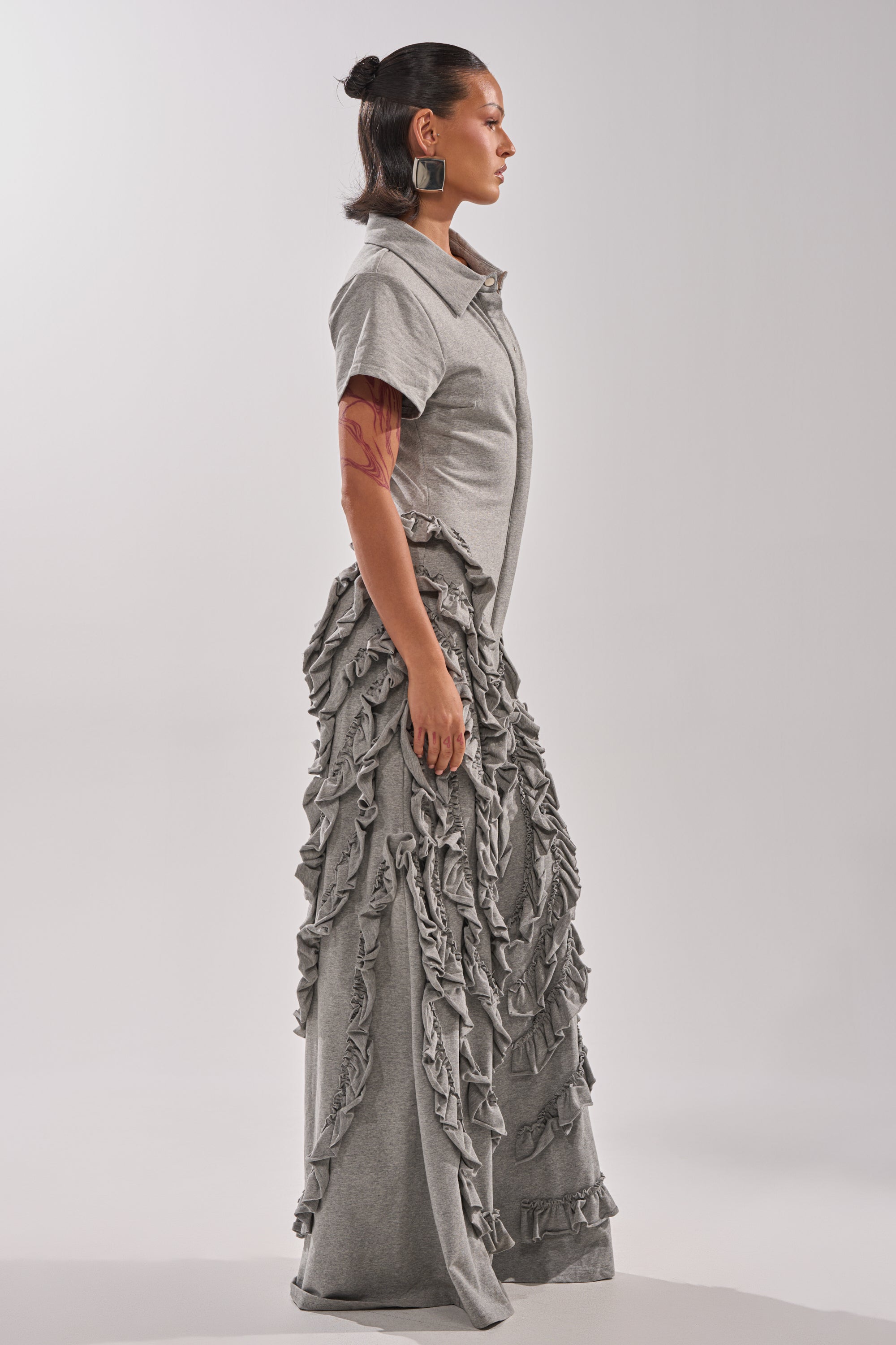A person with dark hair pulled back stands in profile, wearing the LEAH RUFFLE JUMPSUIT—a light gray heathered knit design with wide legs and textured ruffled details. They have a visible arm tattoo and large, square earrings.