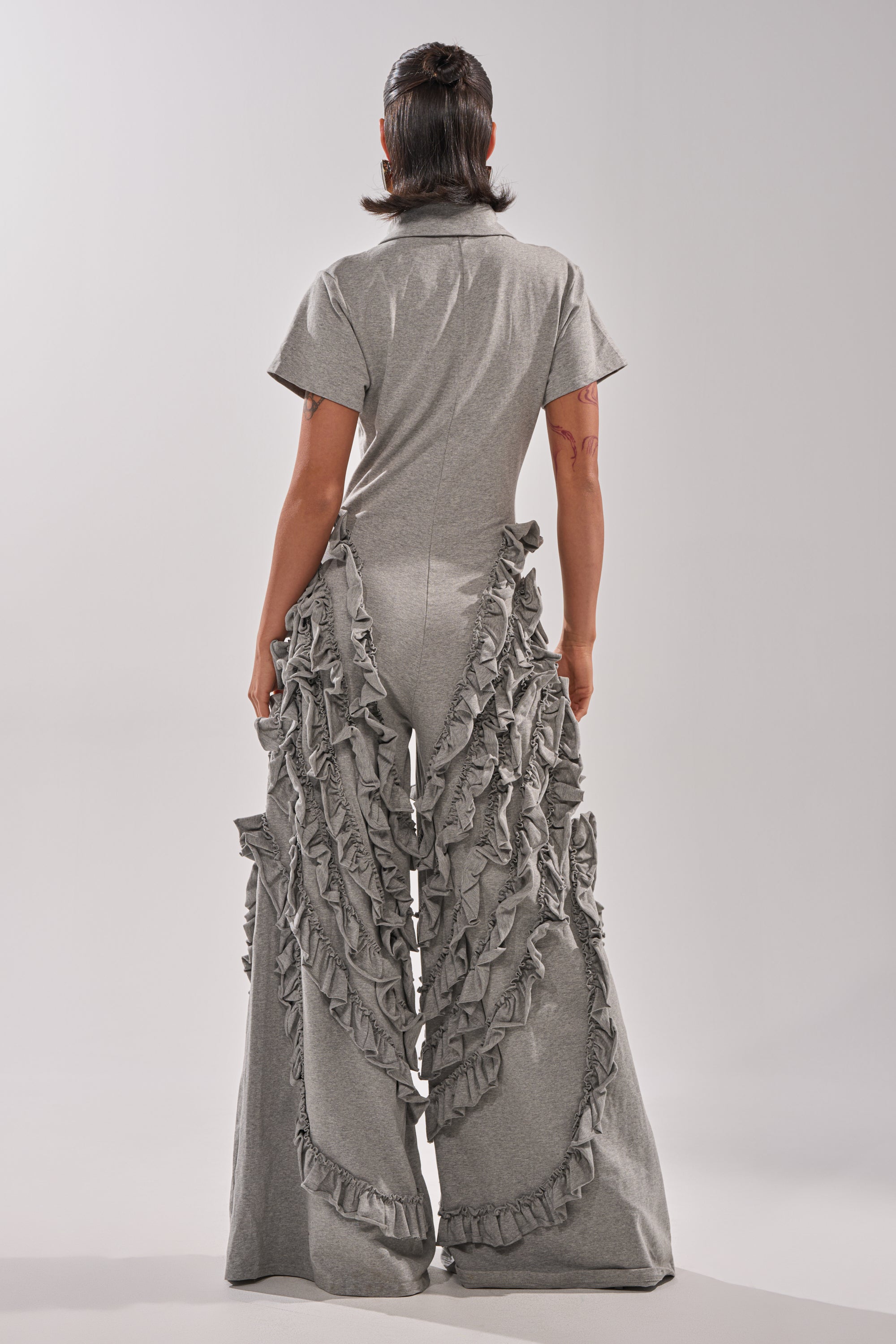 A person with dark hair pulled back stands facing away in the LEAH RUFFLE JUMPSUIT, featuring wide legs and cascading ruffles down the back, against a plain light-colored background.