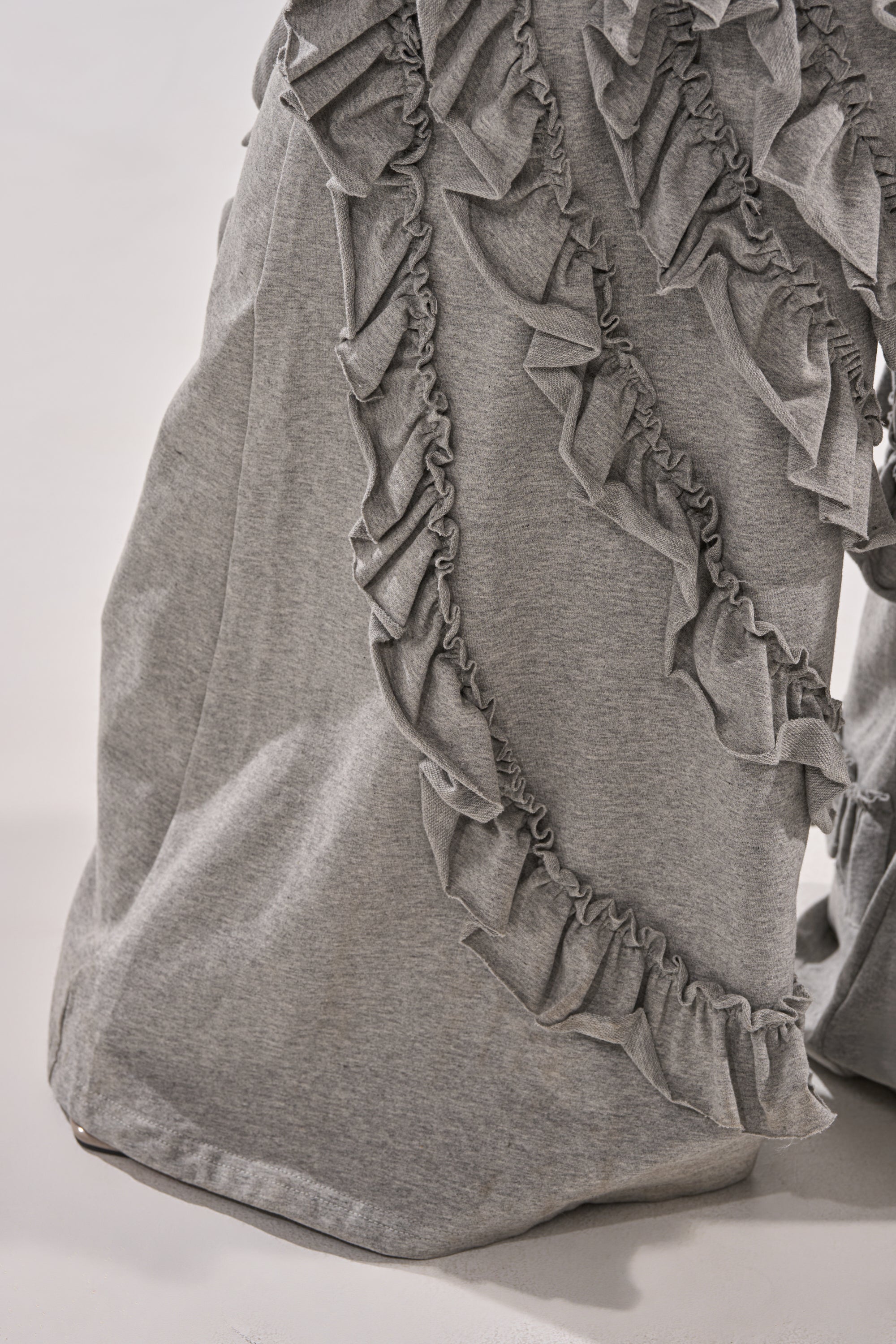 A close-up of the LEAH RUFFLE JUMPSUIT in gray, featuring cascading ruffle seams that create a wavy, textured pattern. The soft fabric drapes naturally for an elegant look.