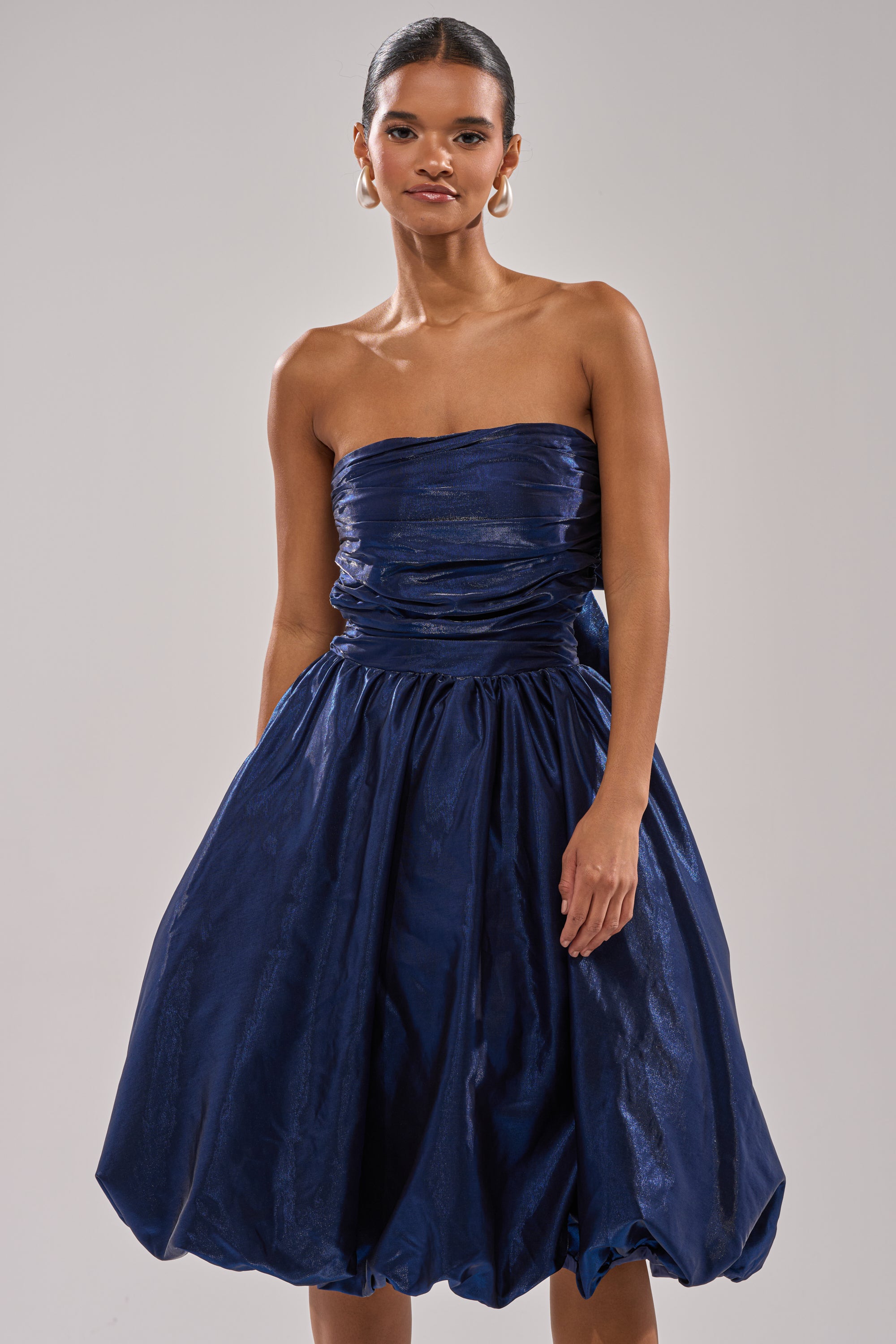 A woman models the LUXE LIFE SATIN MIDI DRESS, a strapless navy blue midi featuring a ruched bodice and voluminous shiny skirt. Her hair is pulled back and she accessorizes with large hoop earrings. 0