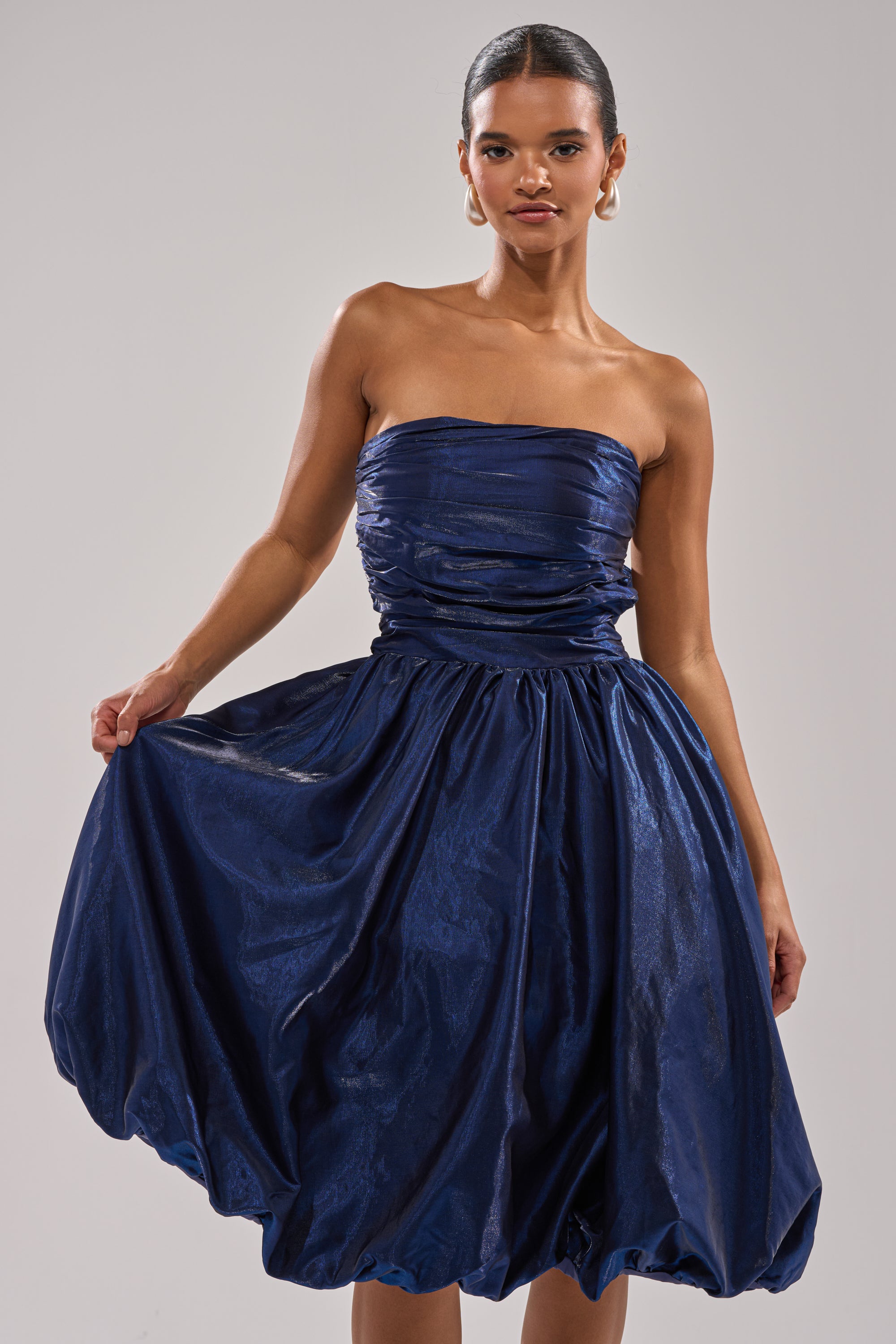 A woman stands against a plain light background, holding out the skirt of the LUXE LIFE SATIN MIDI DRESS—a strapless, ruched deep blue satin cocktail dress with a full skirt. 1
