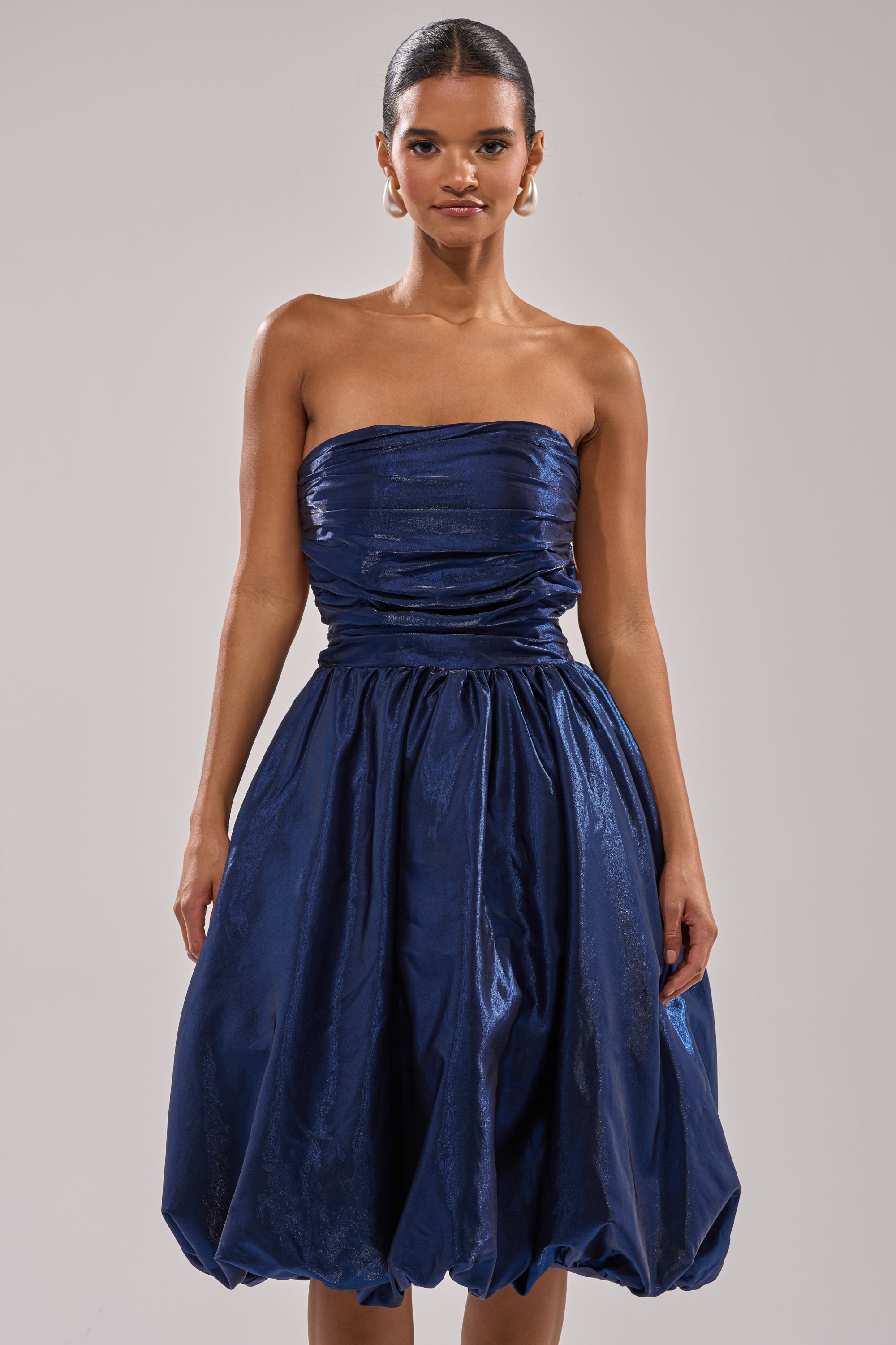 A woman stands confidently against a plain light background wearing the LUXE LIFE SATIN MIDI DRESS—a strapless, dark blue cocktail dress featuring ruched details and a voluminous bubble skirt. 2