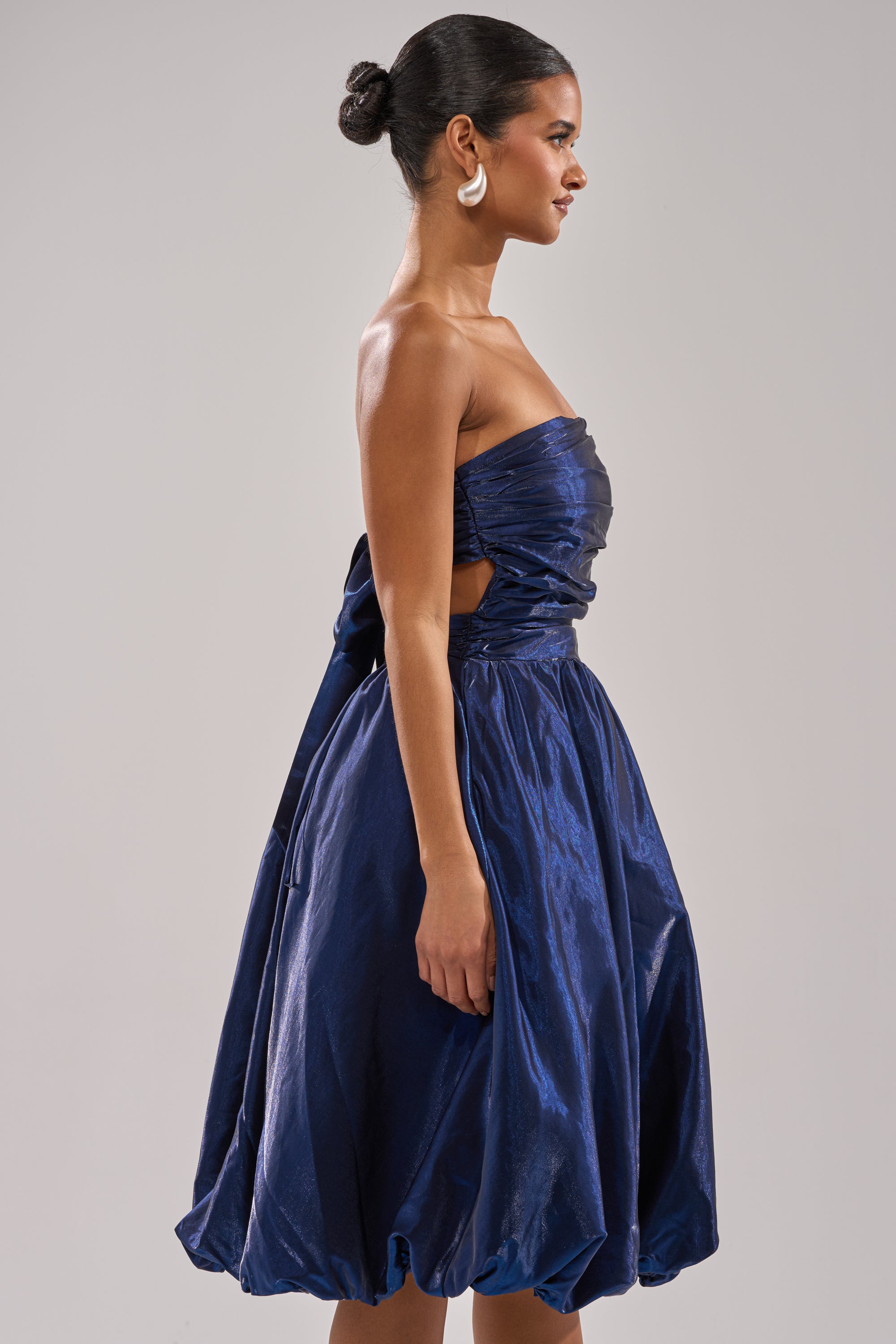 A woman with dark hair in a bun stands in profile against a plain light gray background, wearing the LUXE LIFE SATIN MIDI DRESS—strapless, shiny dark blue satin with a voluminous skirt, ruched details, and side cutouts. 3
