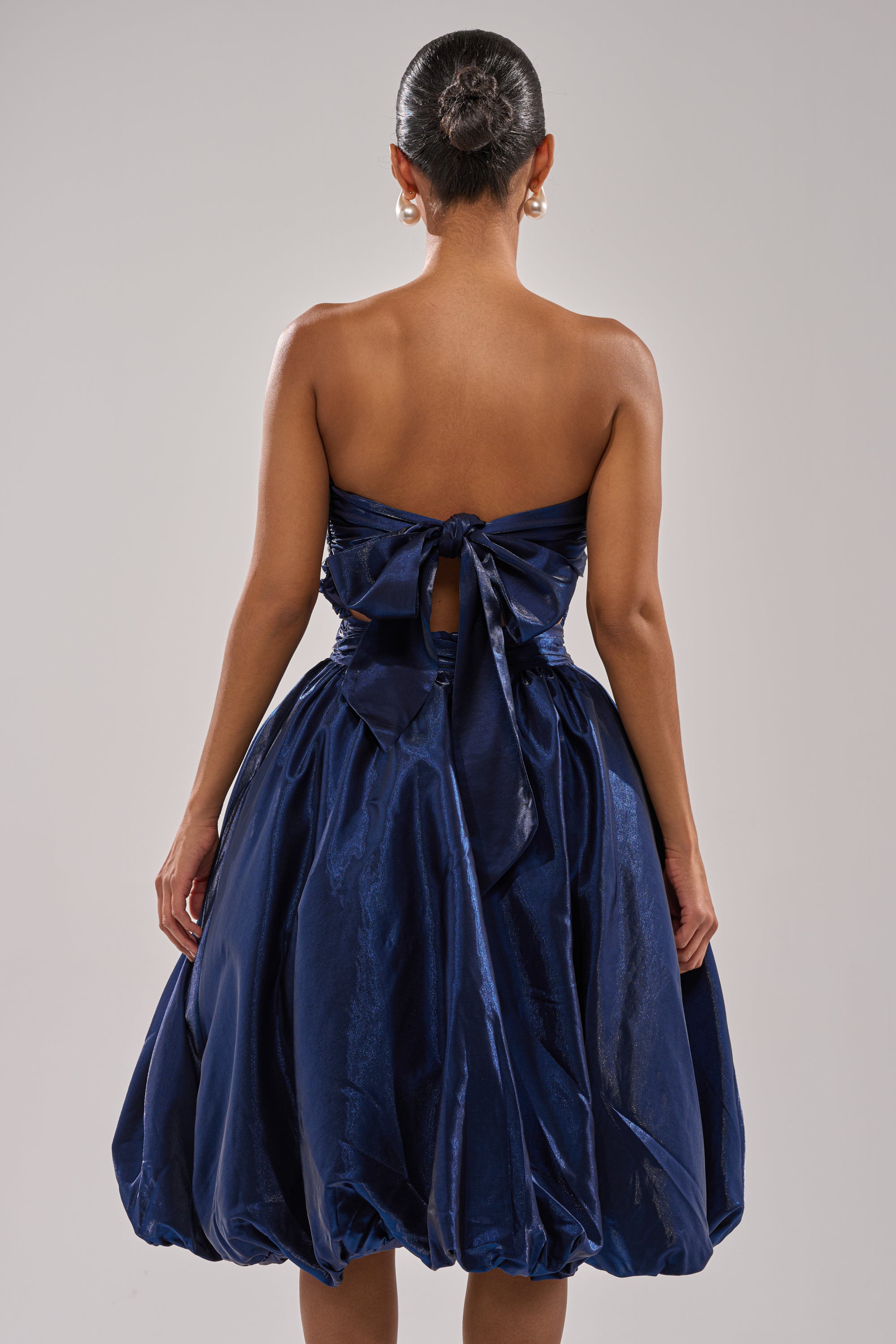 A woman with her hair in a bun models the LUXE LIFE SATIN MIDI DRESS—a strapless, navy blue cocktail dress with a large back bow, voluminous skirt, open back, and elegant ruched construction—while facing away. 4