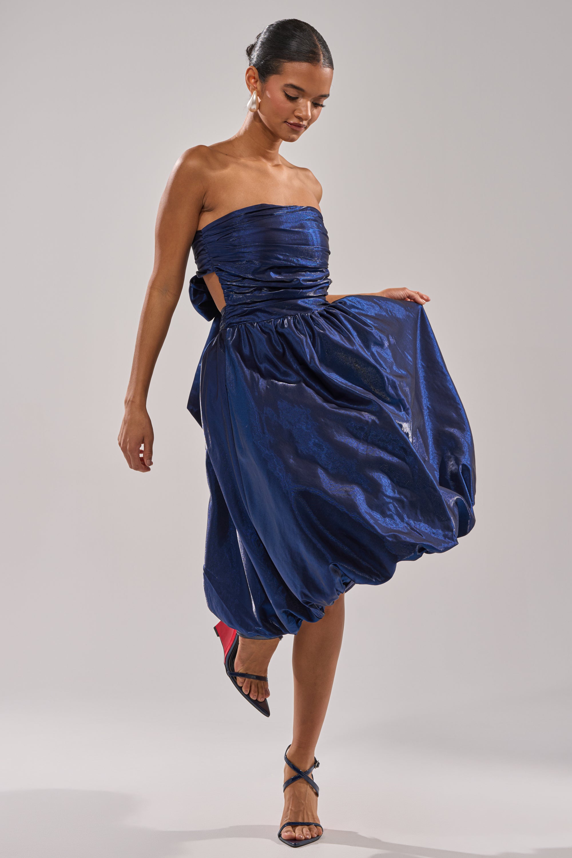 A woman models the LUXE LIFE SATIN MIDI DRESS—a strapless, shiny dark blue bubble-hem midi—posing gracefully with one side of the skirt held out. She pairs it with strappy high-heeled sandals and a sleek bun hairstyle. 5